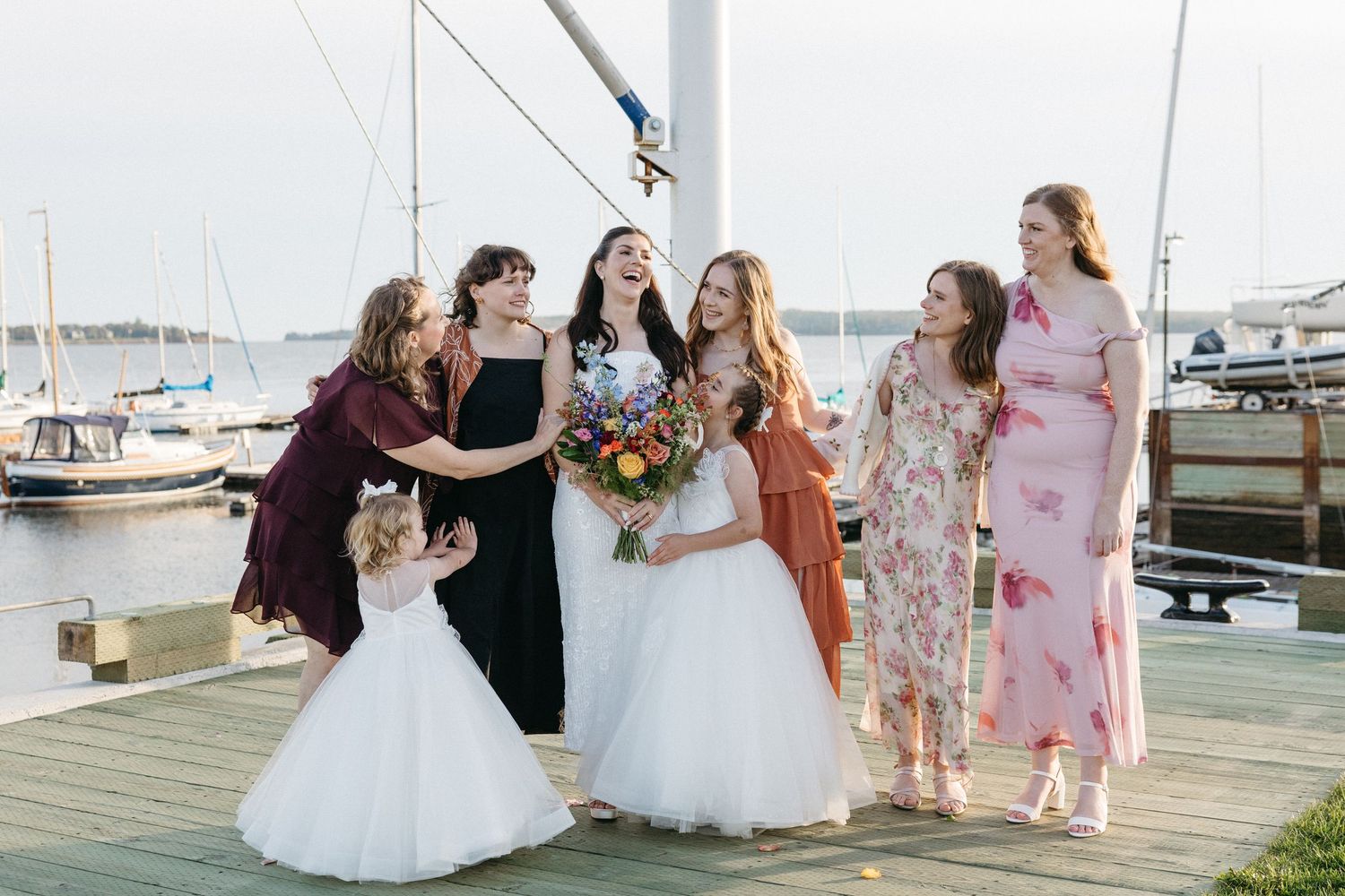 Bride in a white gown holding a colorful bouquet laughs with bridesmaids and two flower girls on a sunny waterfront dock with sailboats.