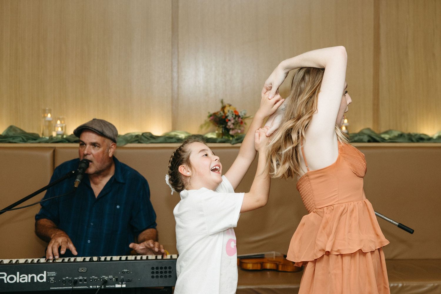 A young girl in a white shirt laughs while dancing with a woman in an orange dress as a keyboardist in a cap plays at a festive event.