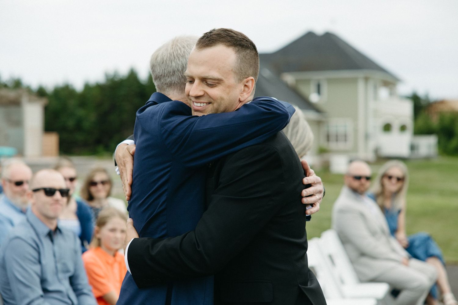 Groom smiling while sharing warm hug with guest at outdoor wedding ceremony.