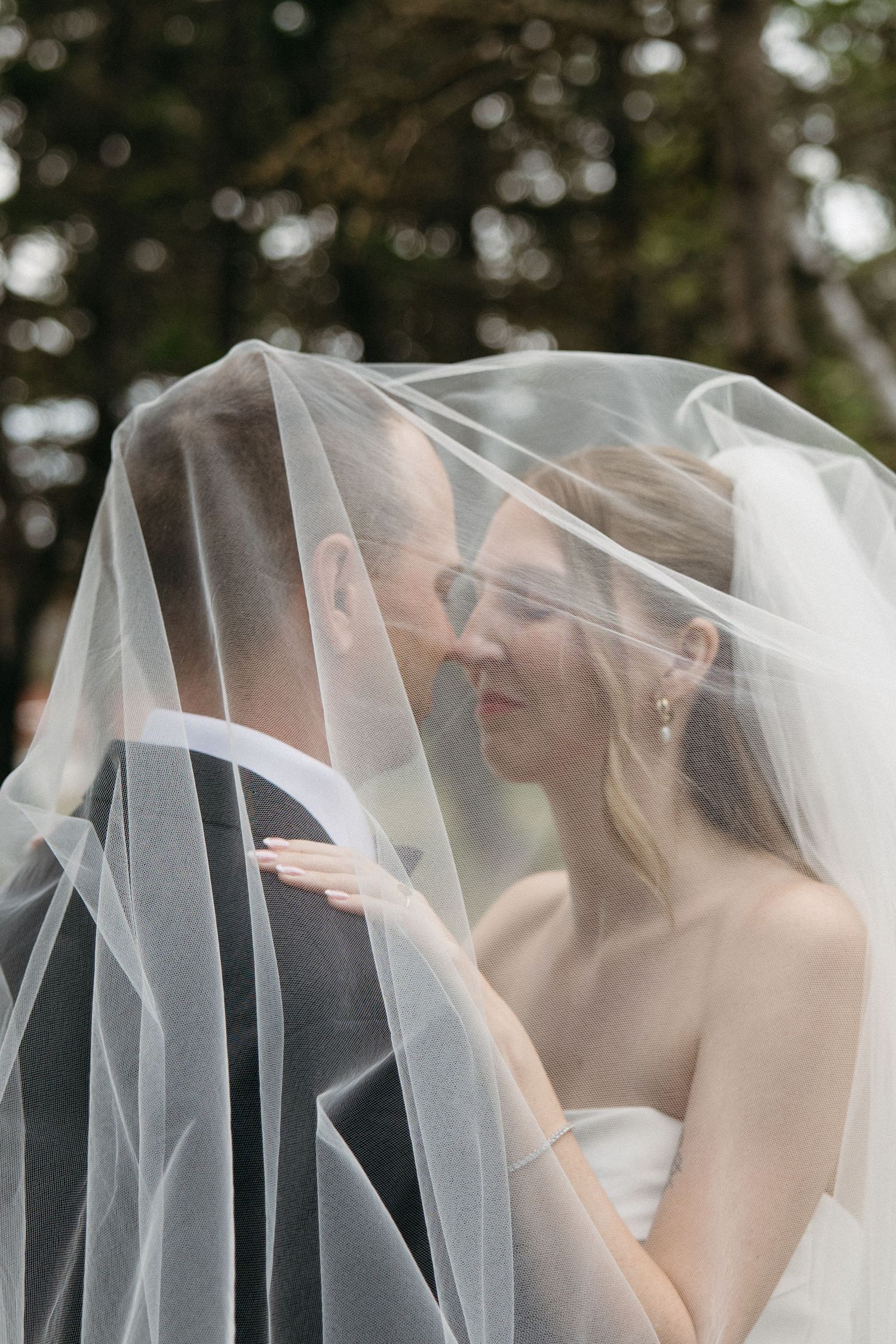 Bride and groom share an intimate moment together beneath a flowing white wedding veil outdoors.