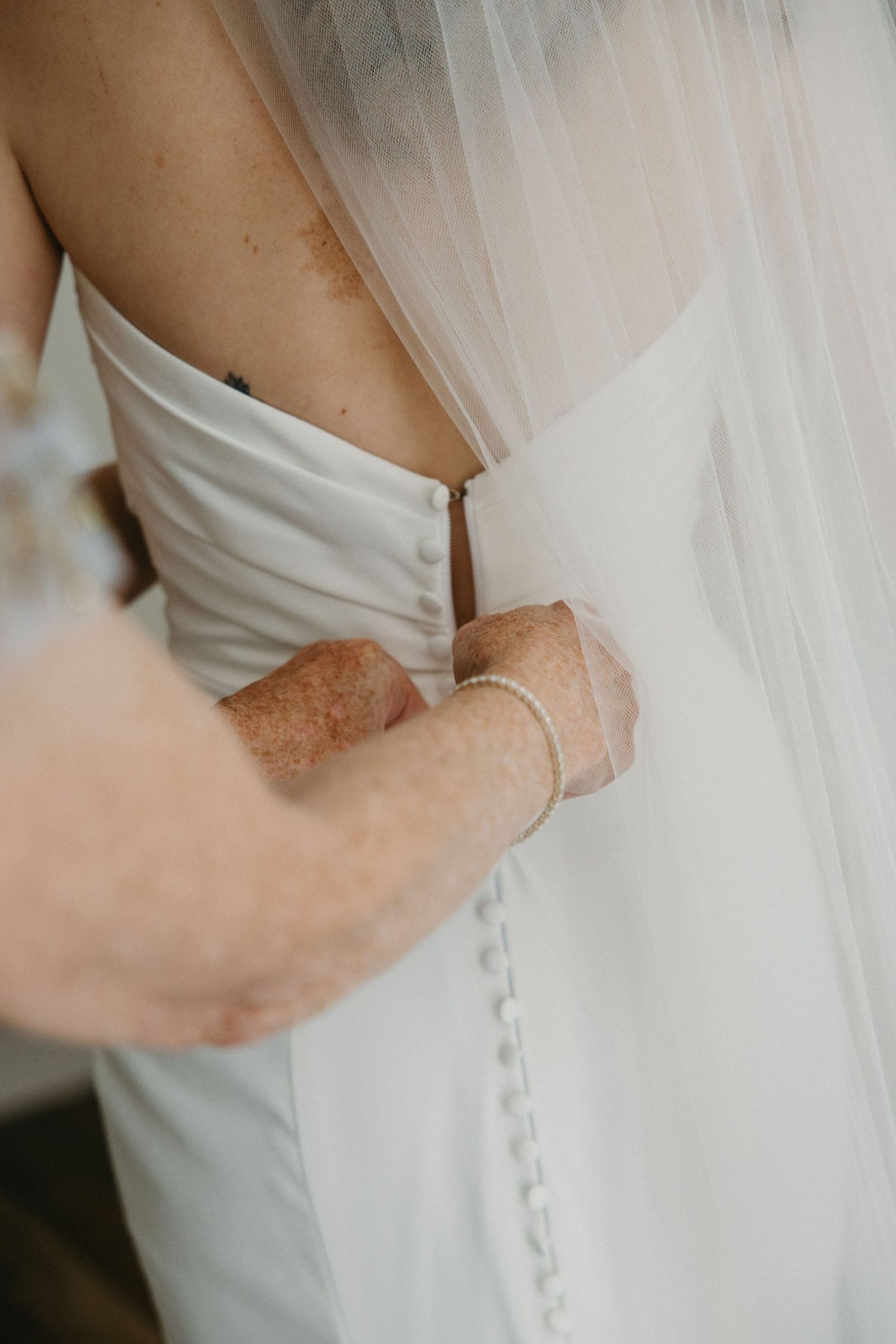 An older woman fastens the back buttons of a bride's white strapless wedding gown.