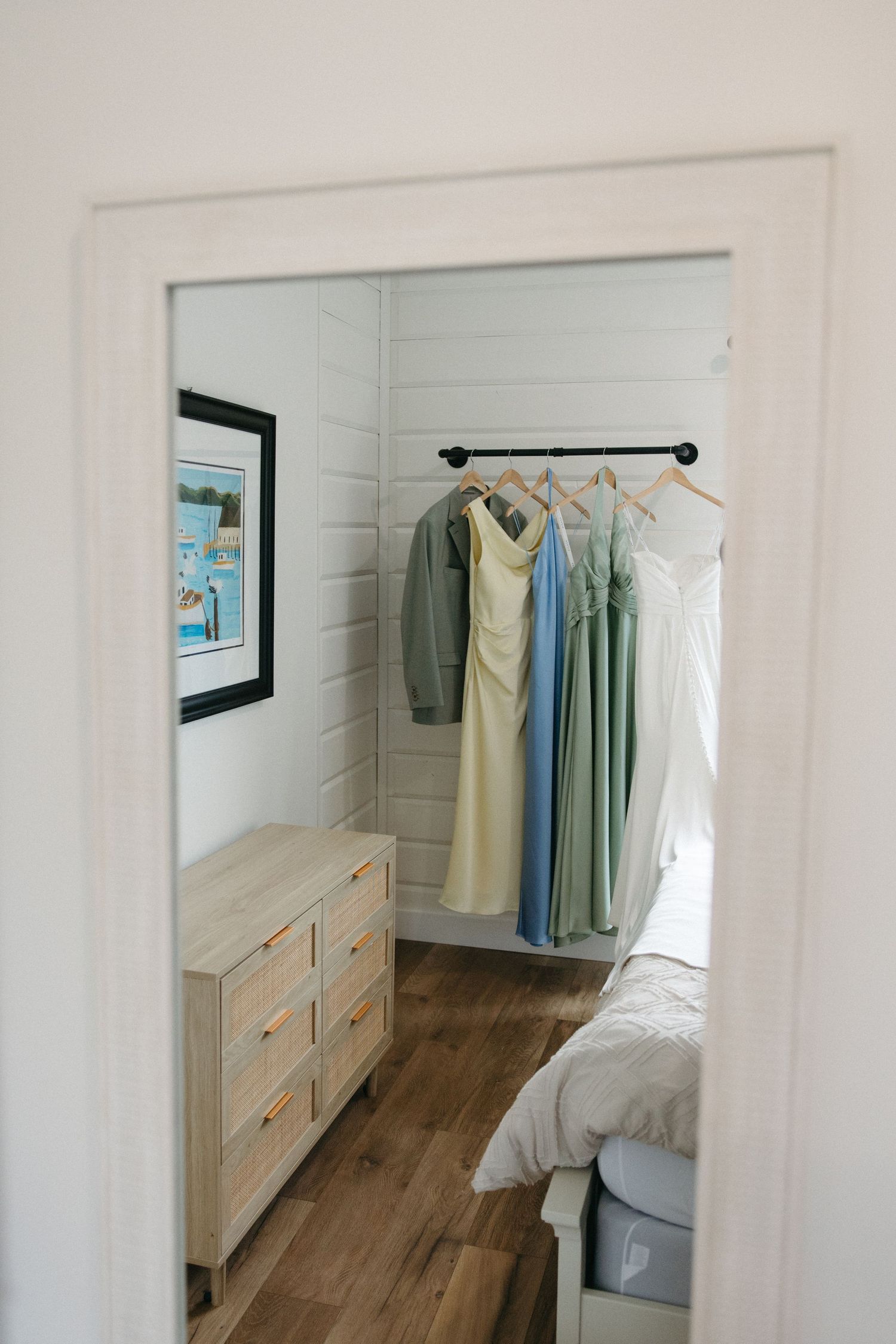 Bridesmaid dresses and wedding gown hanging on a rack in a bright white shiplap bedroom with wood floors.