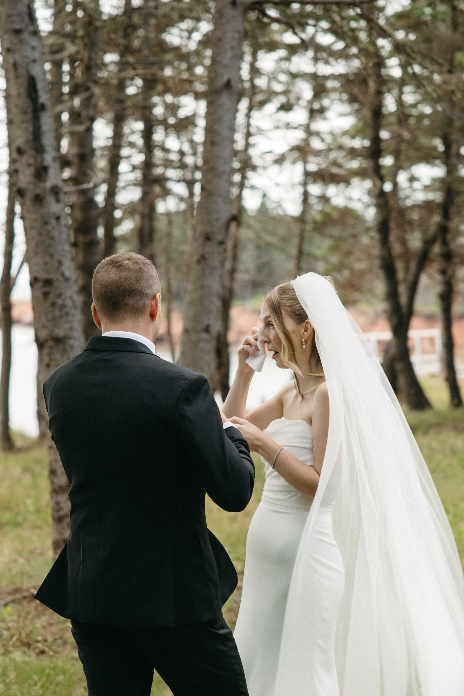Emotional bride in white gown and veil wiping tears during first look with groom in forest setting.