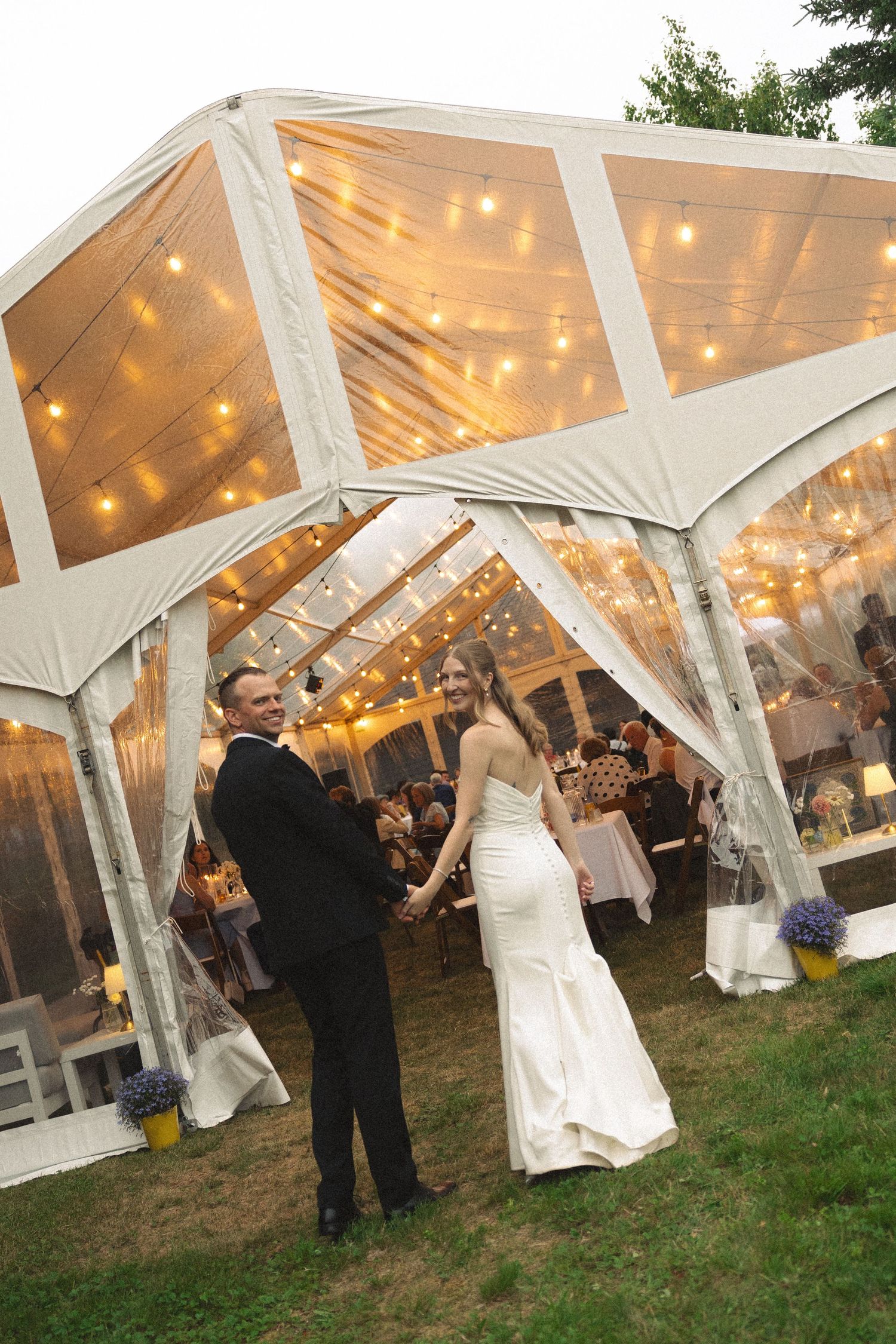Bride and groom hold hands smiling outside illuminated clear wedding tent at dusk.