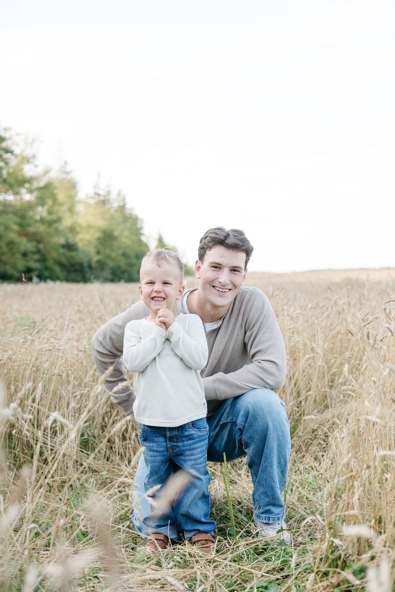 A smiling father crouches beside his young son in a golden wheat field, both wearing casual clothes on a bright day.