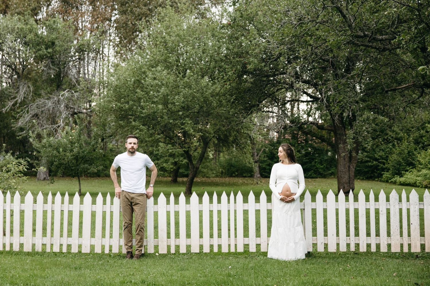 Expecting couple standing apart by white picket fence in green garden, woman in white dress holding baby bump.
