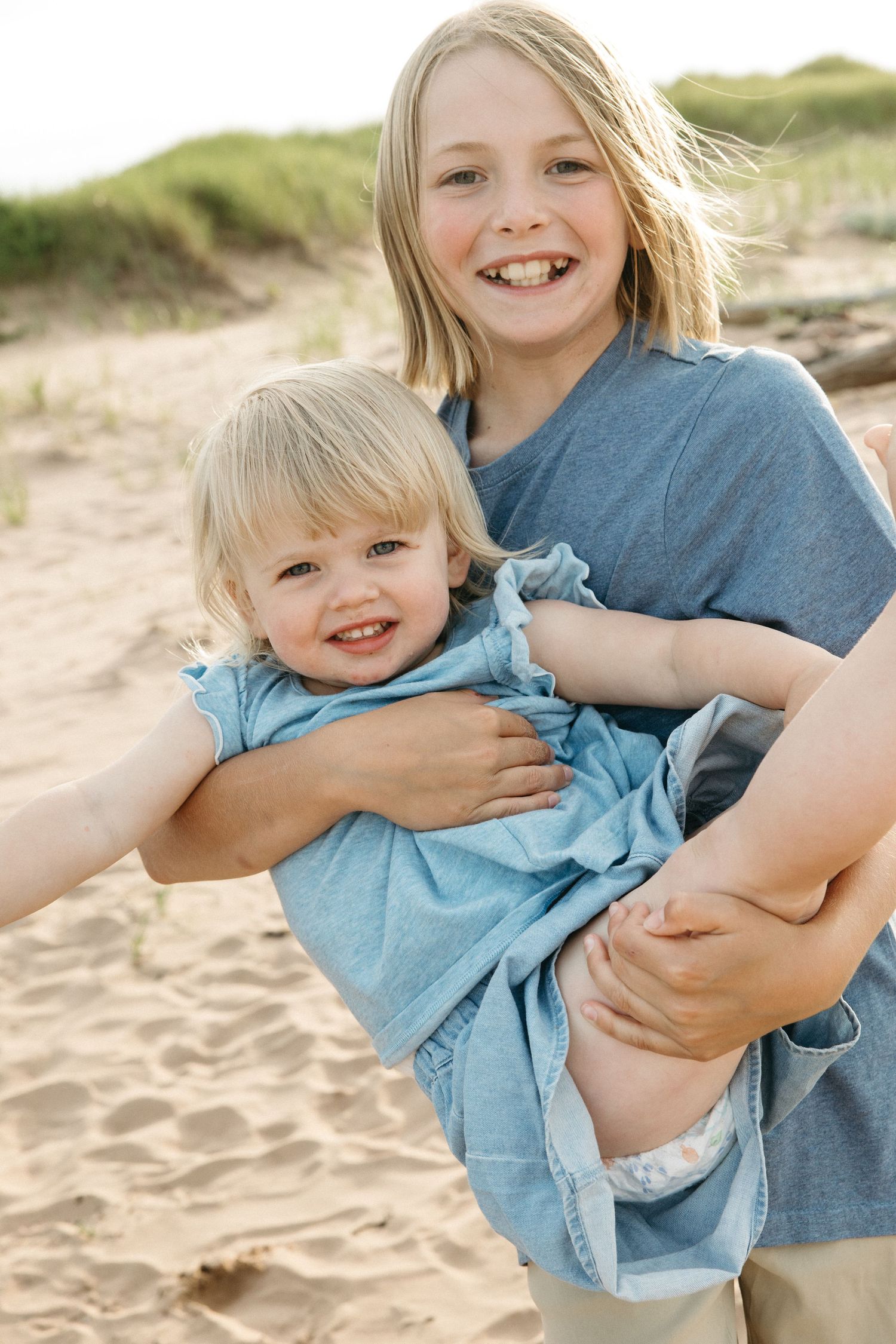 An older blonde child hugs a laughing toddler sibling on a sandy beach with dunes behind them.