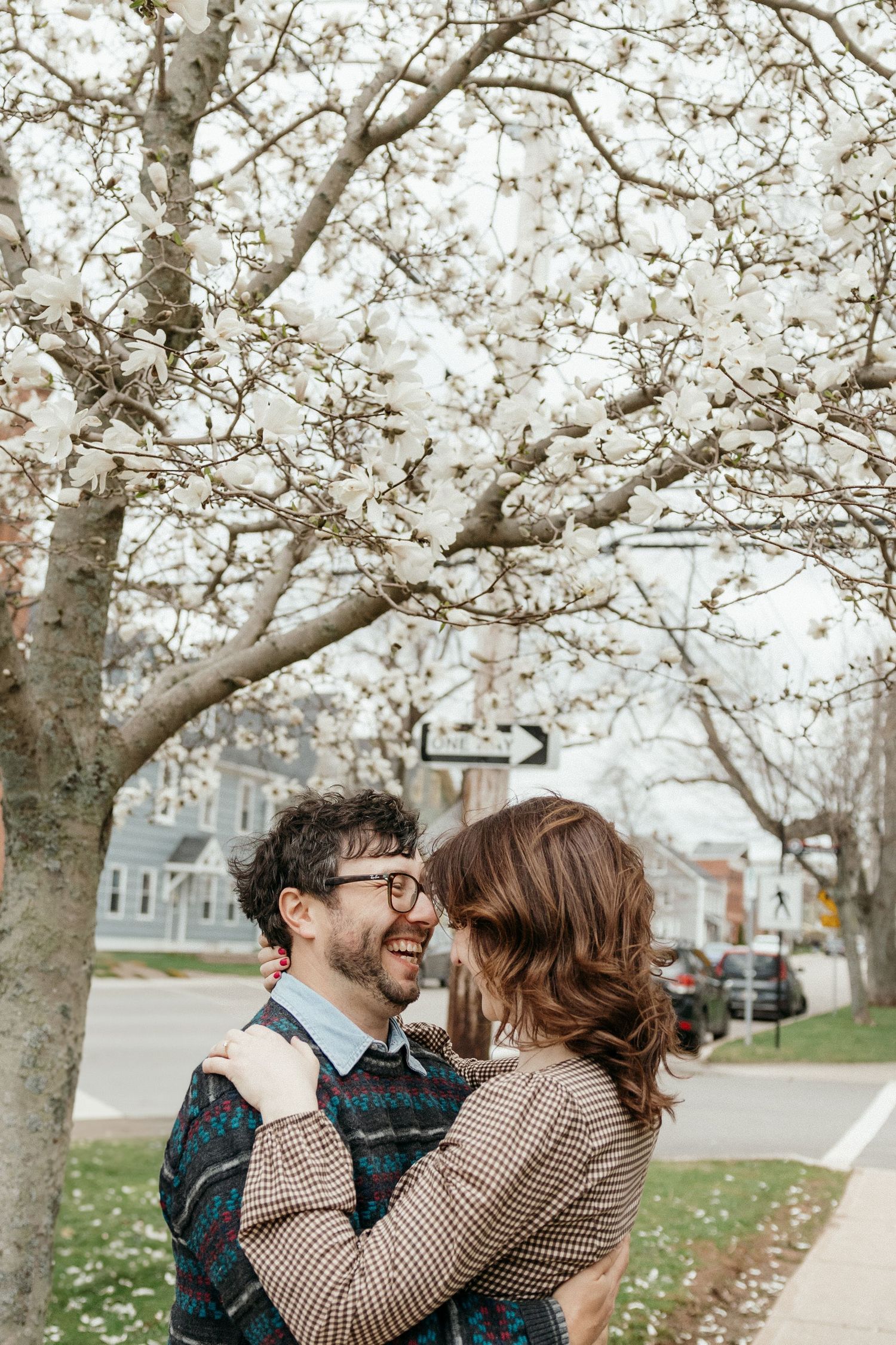 A couple embraces and laughs under a blooming white cherry blossom tree on a sunny spring day.