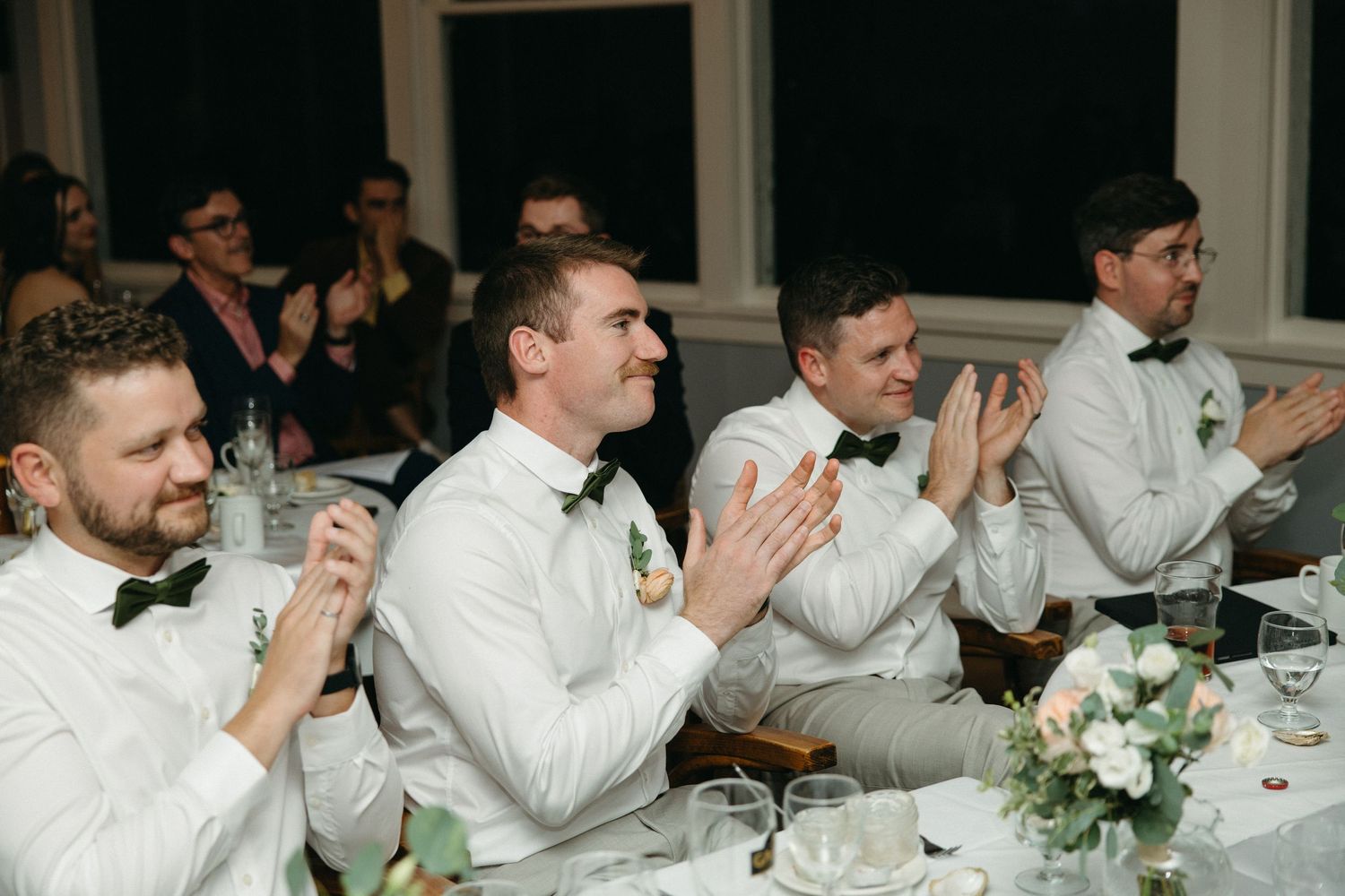 Groomsmen in white suits with green bow ties applaud during wedding reception speeches at elegantly decorated table.