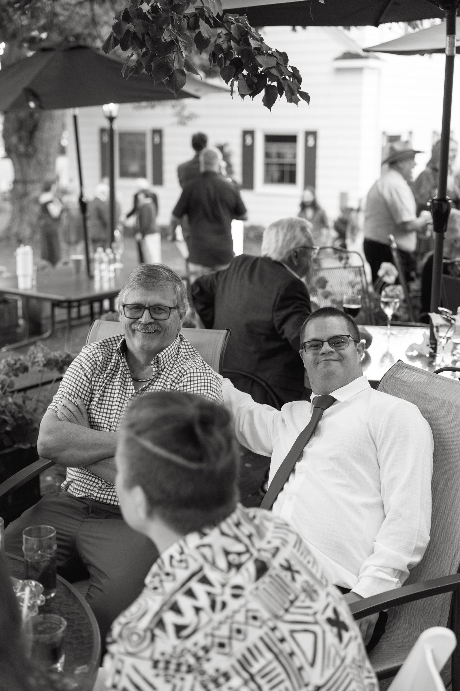 Two men in glasses smile and relax at an outdoor wedding cocktail hour in black and white photo.