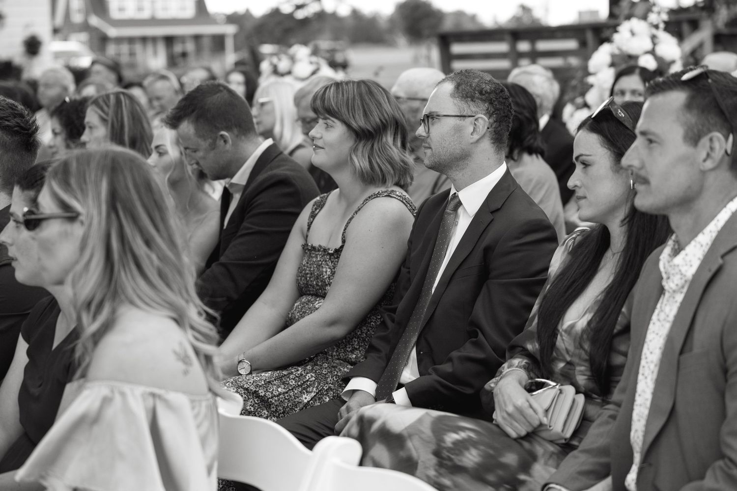 Wedding guests seated outdoors smile during a ceremony in this black and white candid photo.