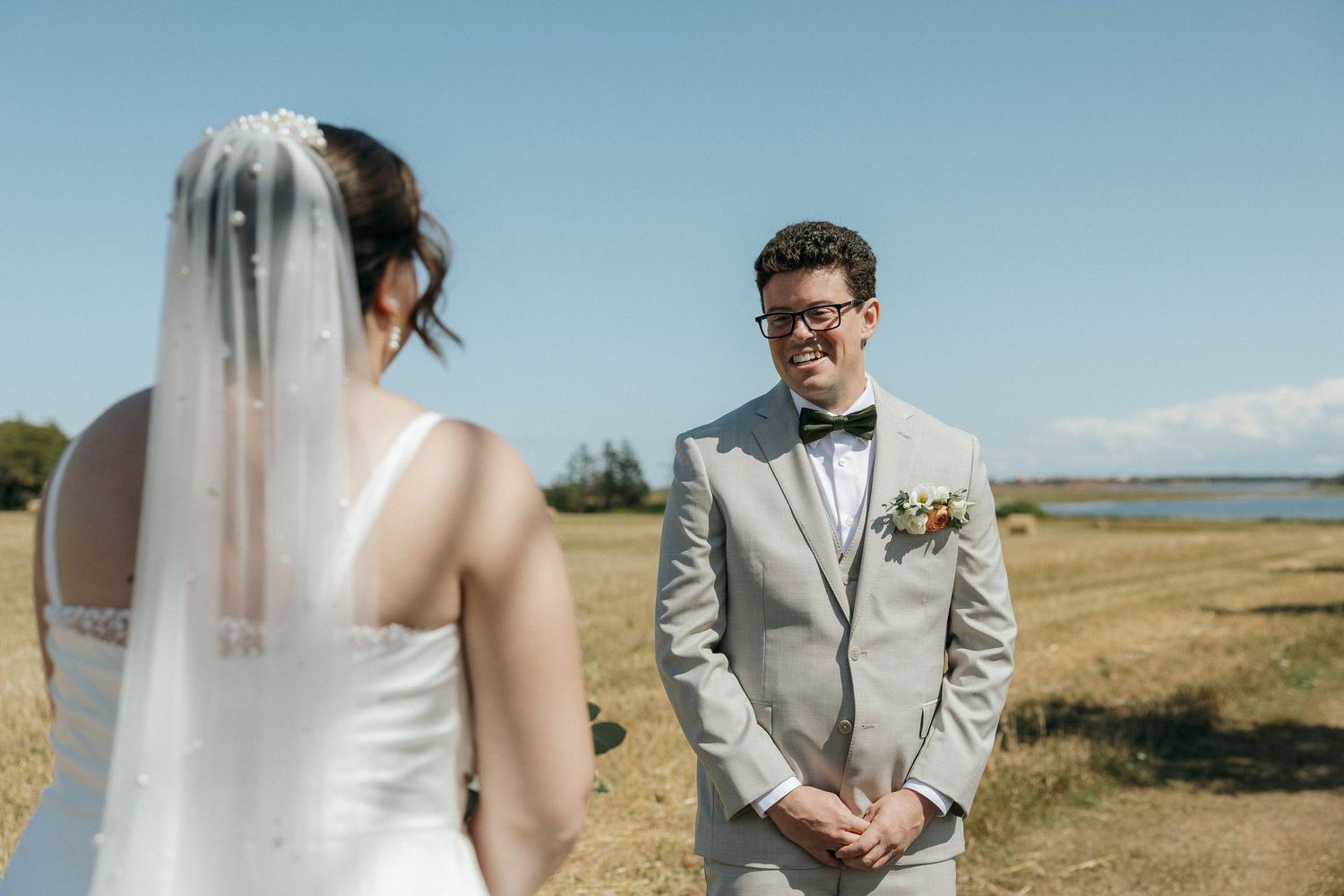 Emotional groom in gray suit smiling at bride during outdoor first look in golden field by the water.