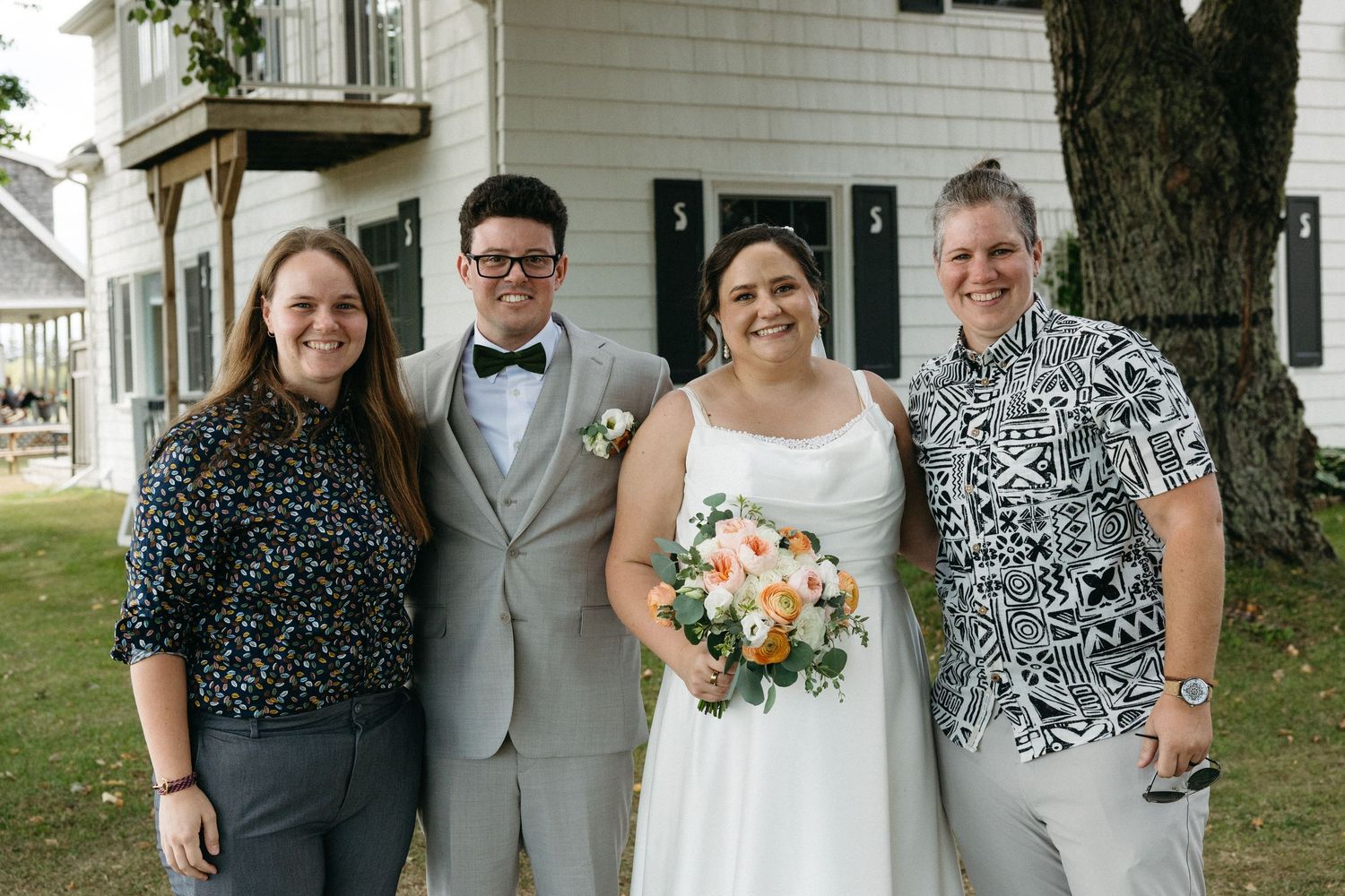 Bride holding bouquet poses with three guests in front of white historic building.