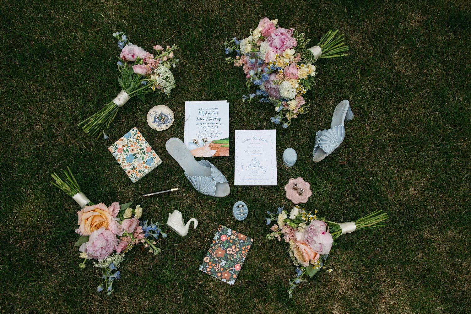 Elegant wedding flat lay with pink and blue floral bouquets, invitations, blue heels, and decorative items on green grass.