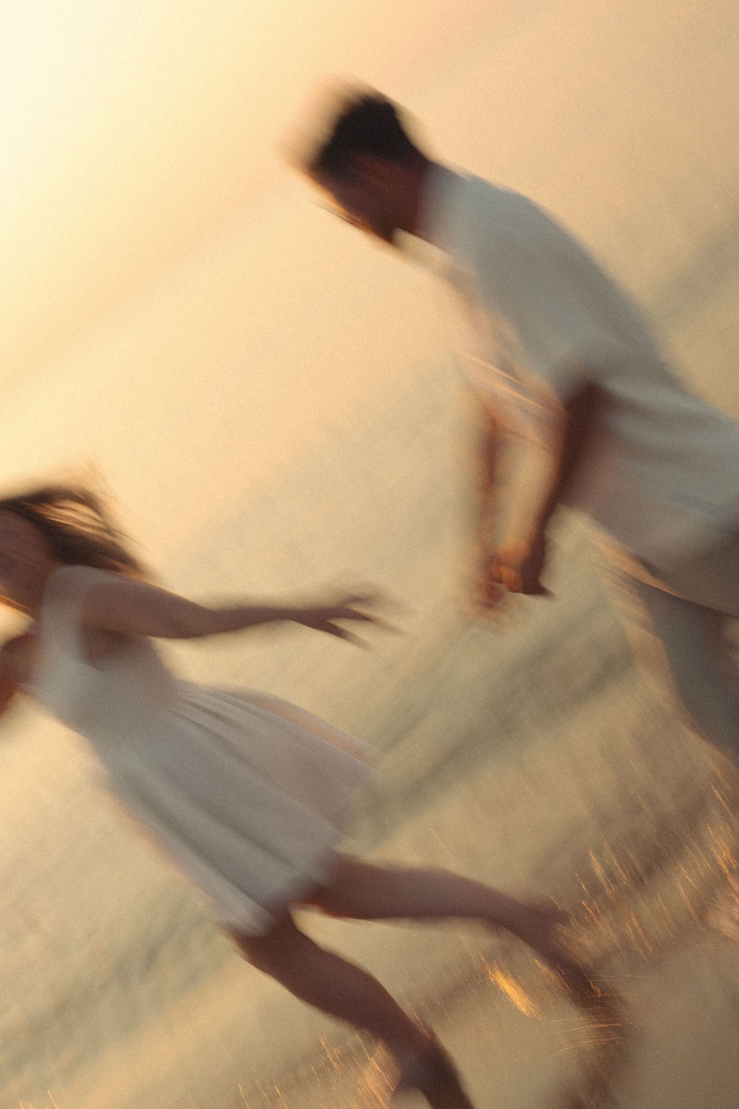 Blurred motion shot of couple in white running playfully along beach at golden sunset.