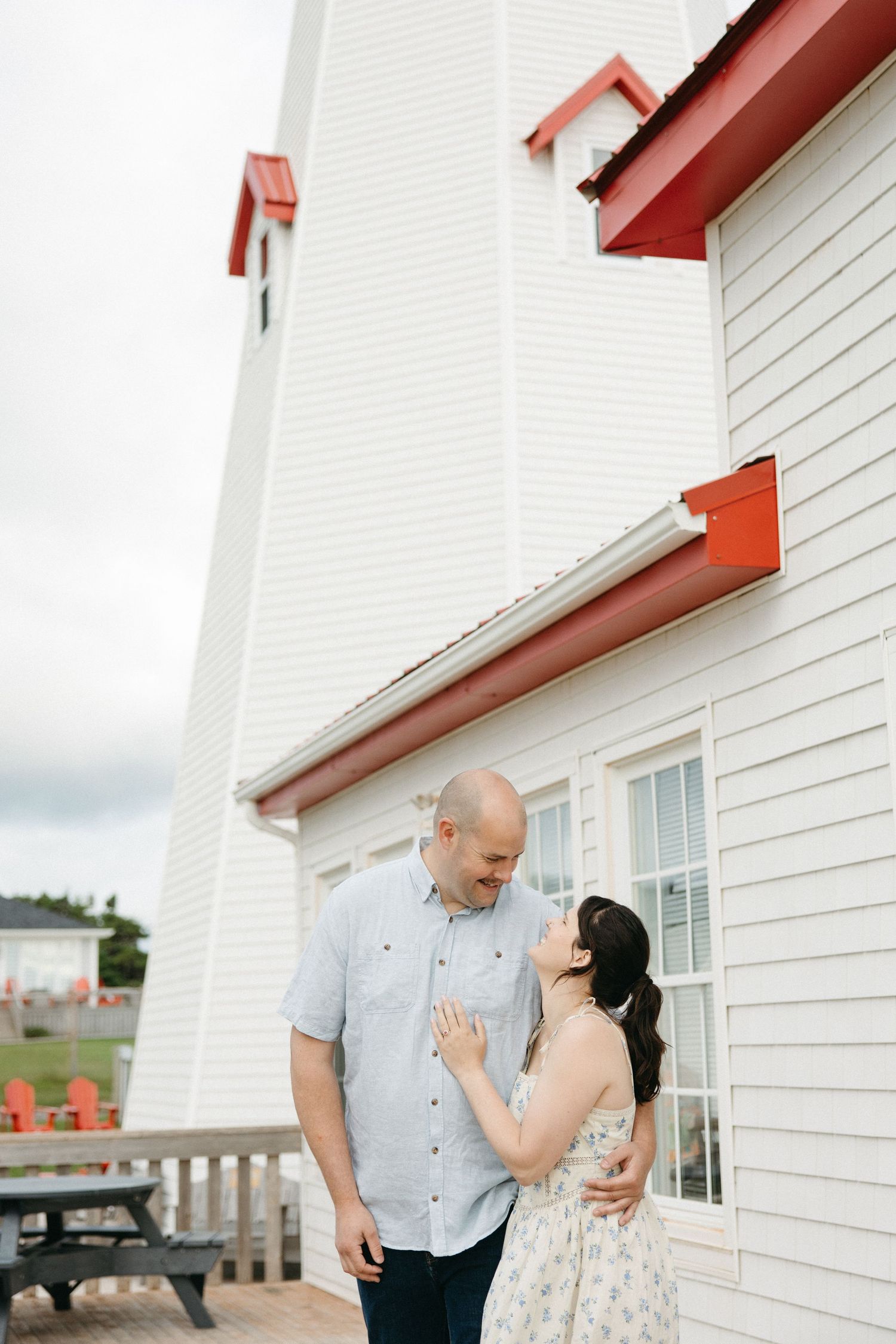 Couple sharing a romantic moment outside a white lighthouse with red trim on a cloudy day.