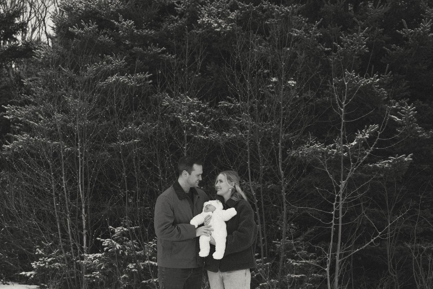A black and white photo of a couple holding their baby in a winter forest setting.