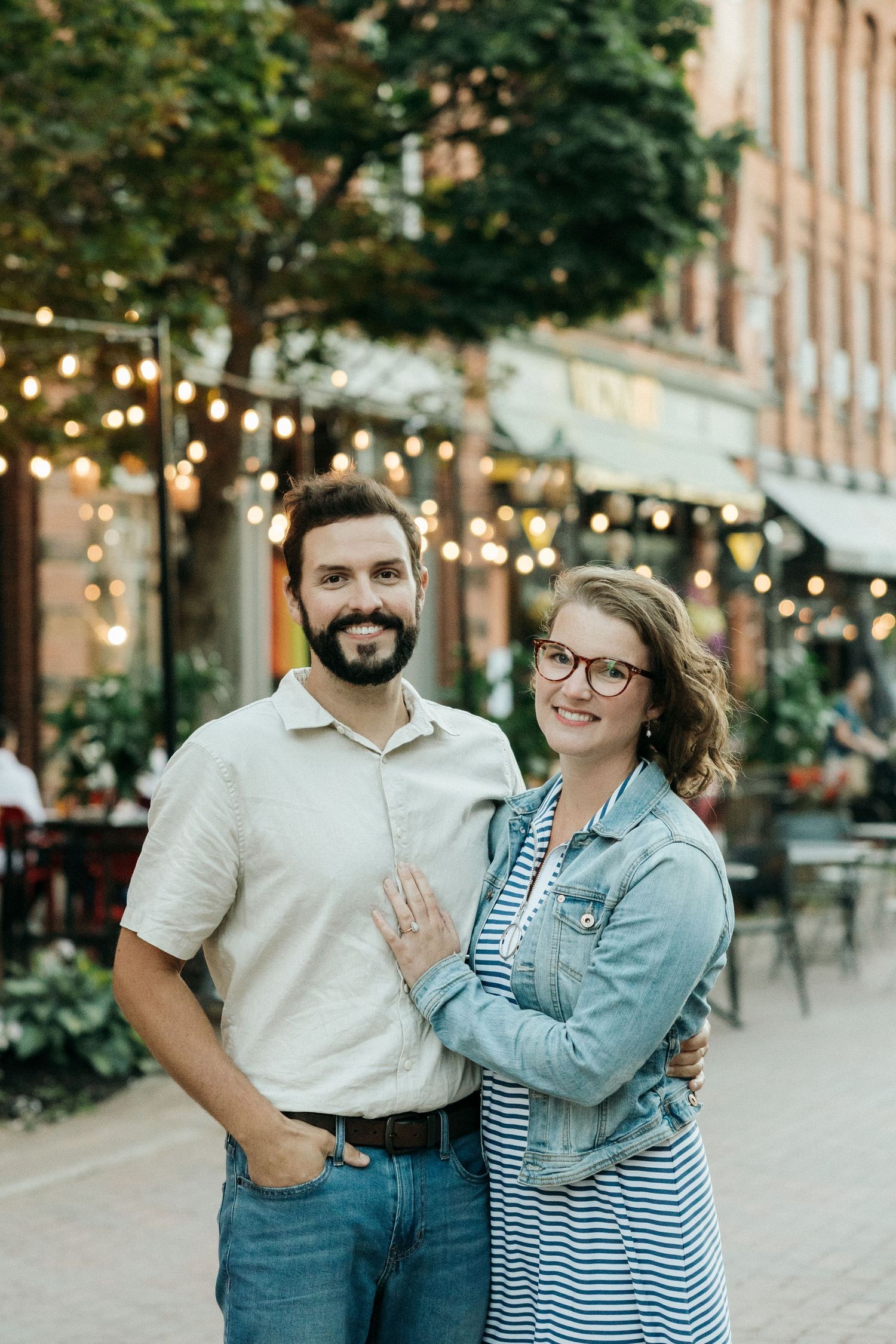 A couple smiles together on a downtown street decorated with string lights and outdoor seating.