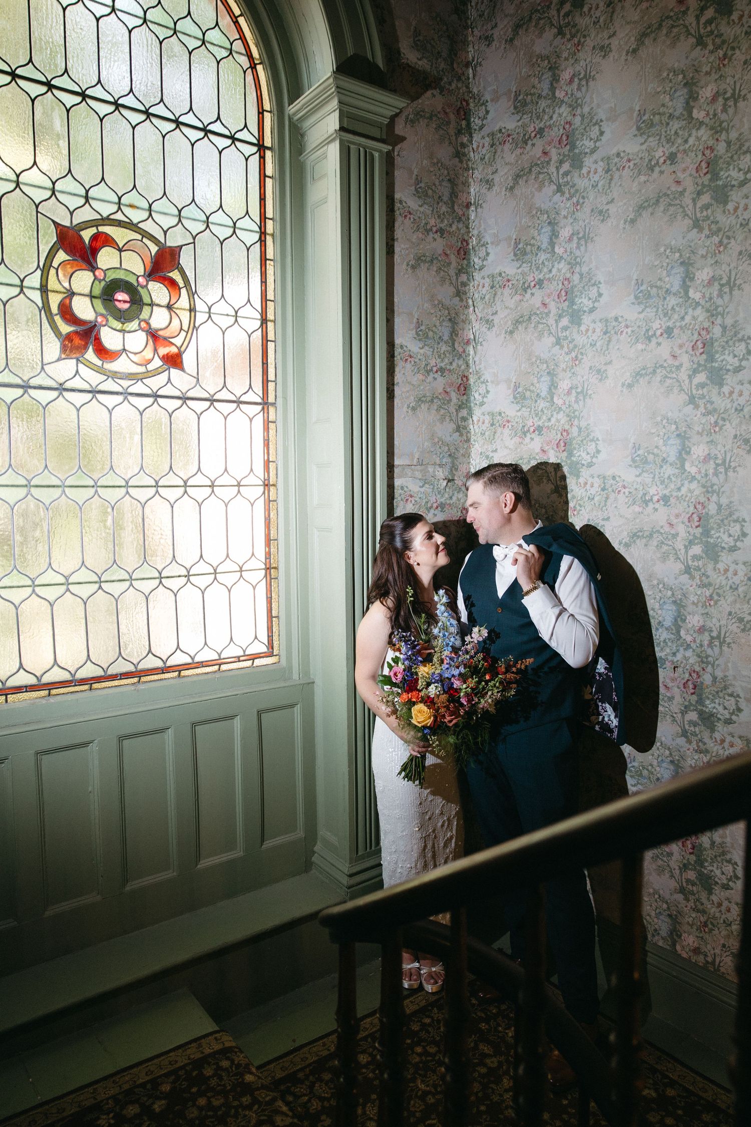 Smiling bride and groom embrace beside a large stained glass window on a vintage staircase with floral wallpaper.