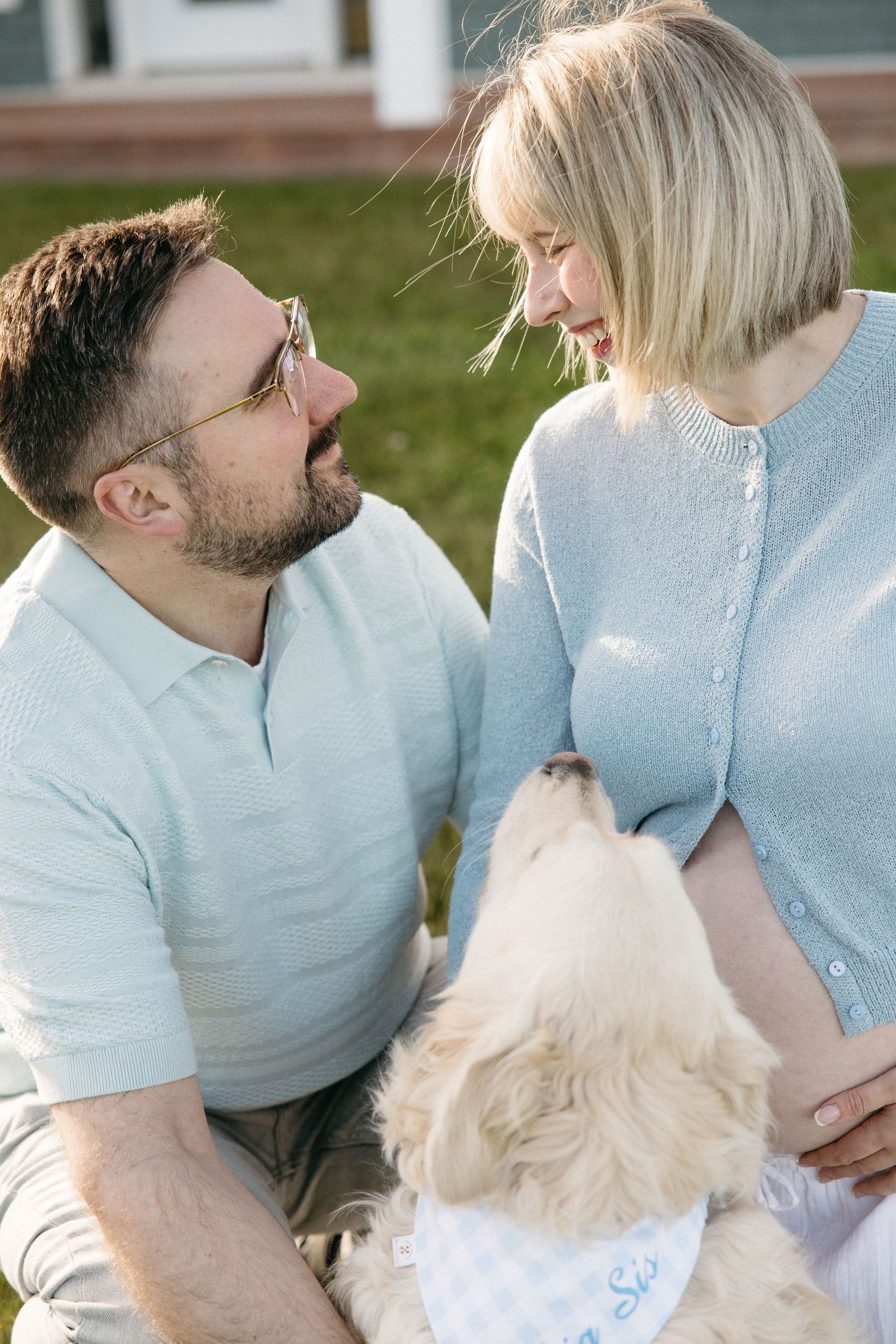 A couple sits outdoors with their white dog, smiling at each other while the woman shows a baby bump.