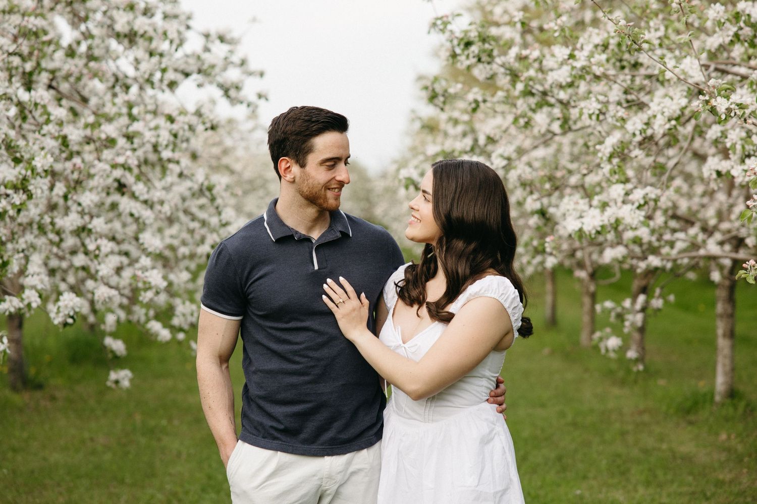 A couple embraces and smiles at each other among rows of white blossoming trees.