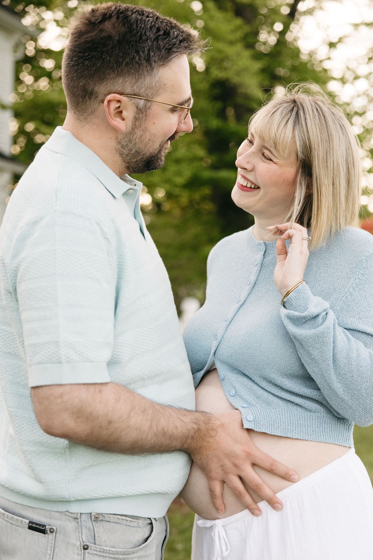 A smiling couple outdoors, the man touching the woman's baby bump, both wearing light blue, sharing a joyful pregnancy moment.
