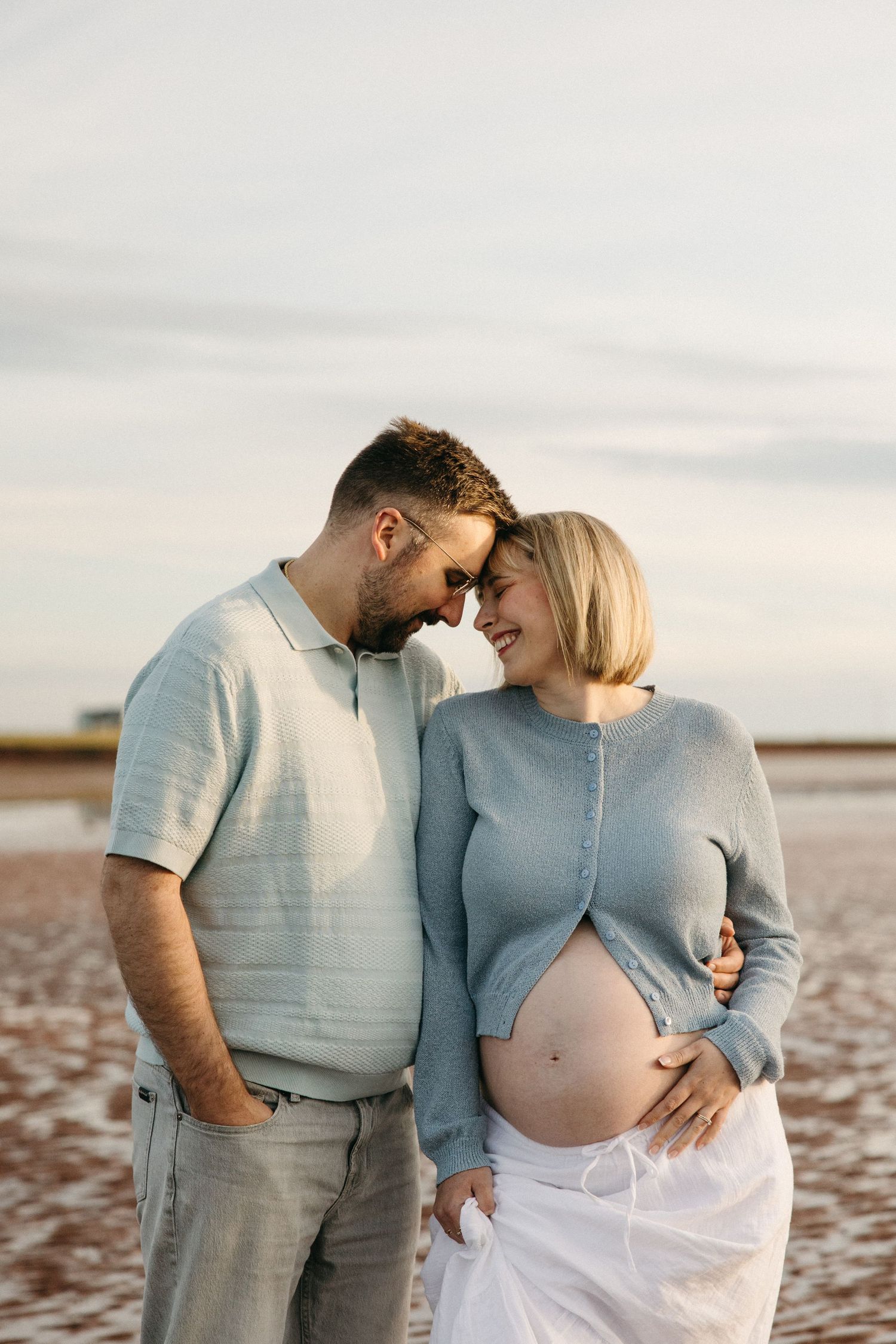Expecting couple touch foreheads romantically on a beach at sunset, man embracing pregnant woman's bump.