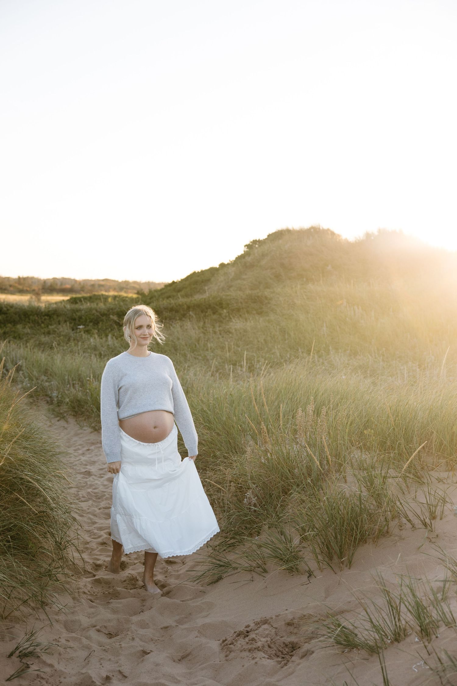 Golden hour maternity photos with sun flare illuminating sand dunes and beach grass.