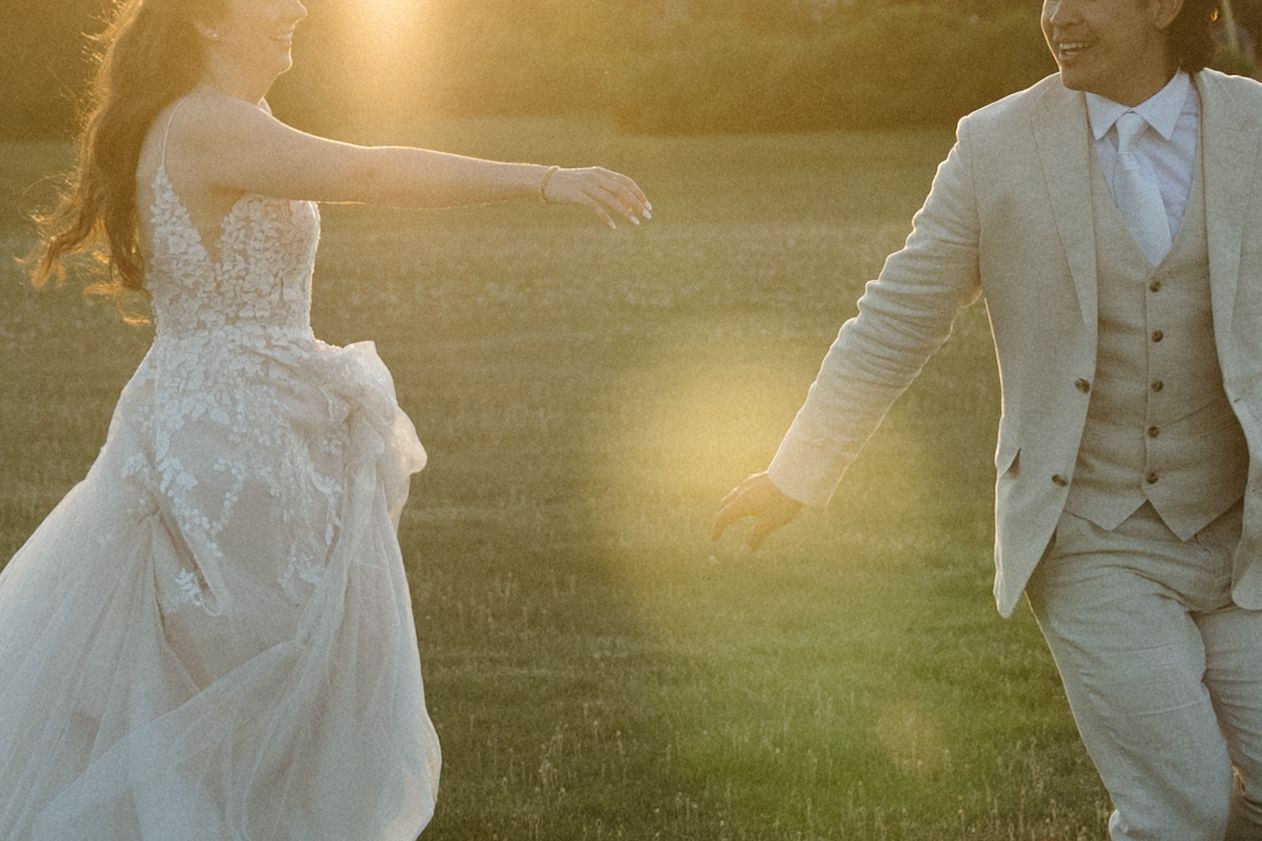 Couple in wedding attire playfully reaching toward each other in golden sunlight.