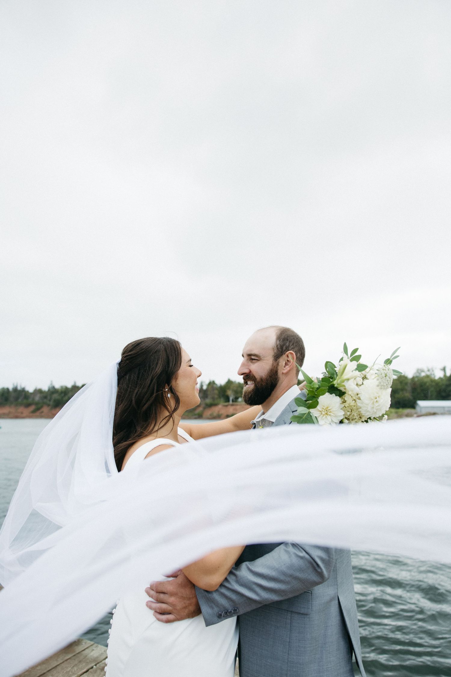 Romantic waterfront moment with flowing white veil against cloudy sky.