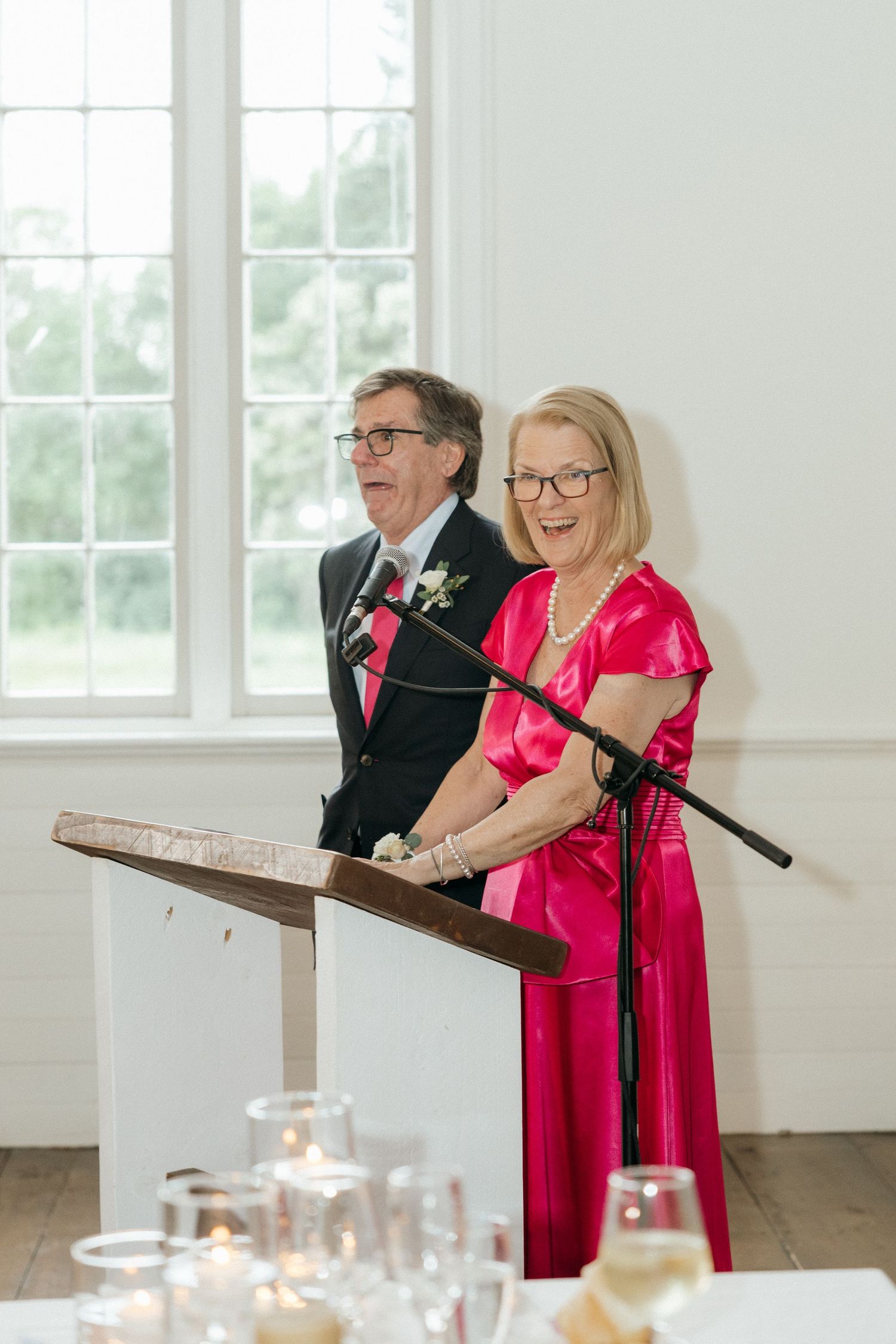 Wedding speakers in bright pink attire deliver speeches at a bright white venue with windows.