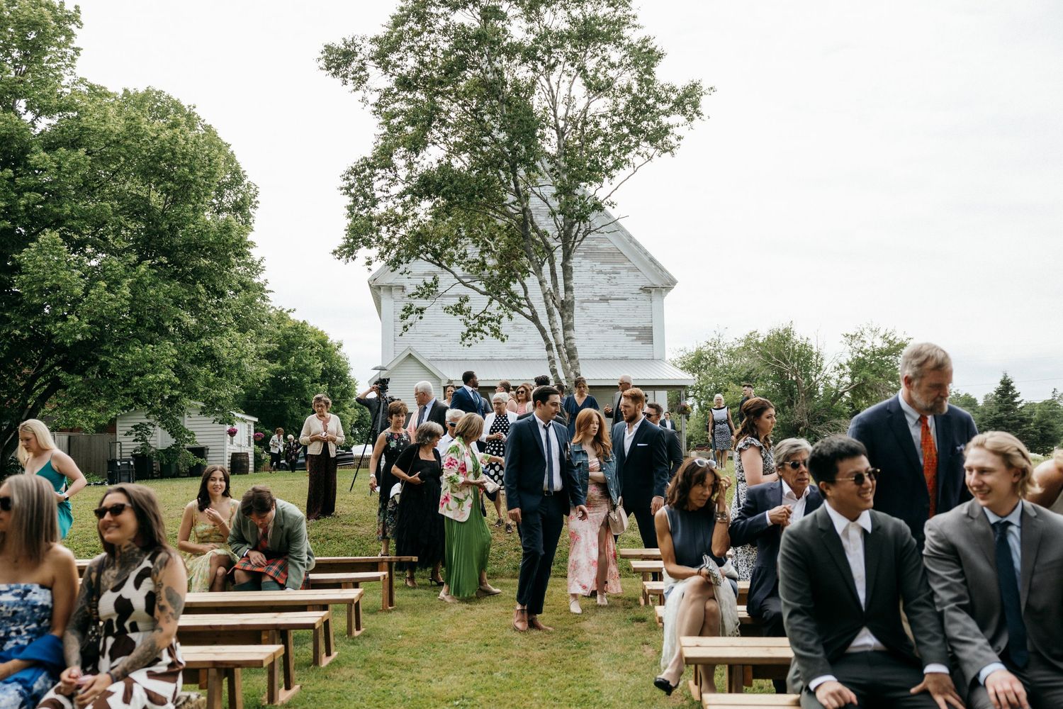 Wedding guests seated on wooden benches await ceremony in front of white church building.
