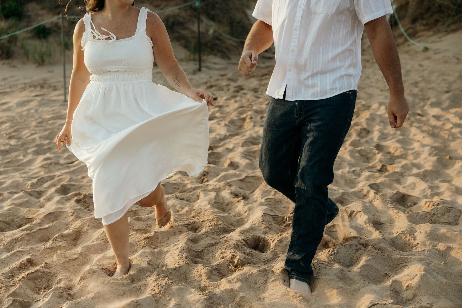 Close-up of feet walking on sandy beach, with flowing white dress and dark pants.