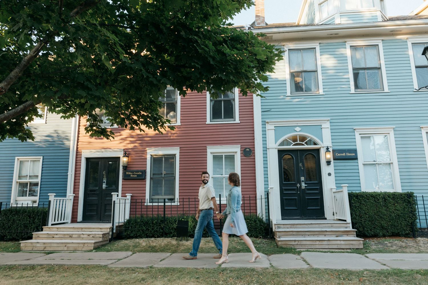 Two people stroll past colorful historic row houses with blue and pink siding.