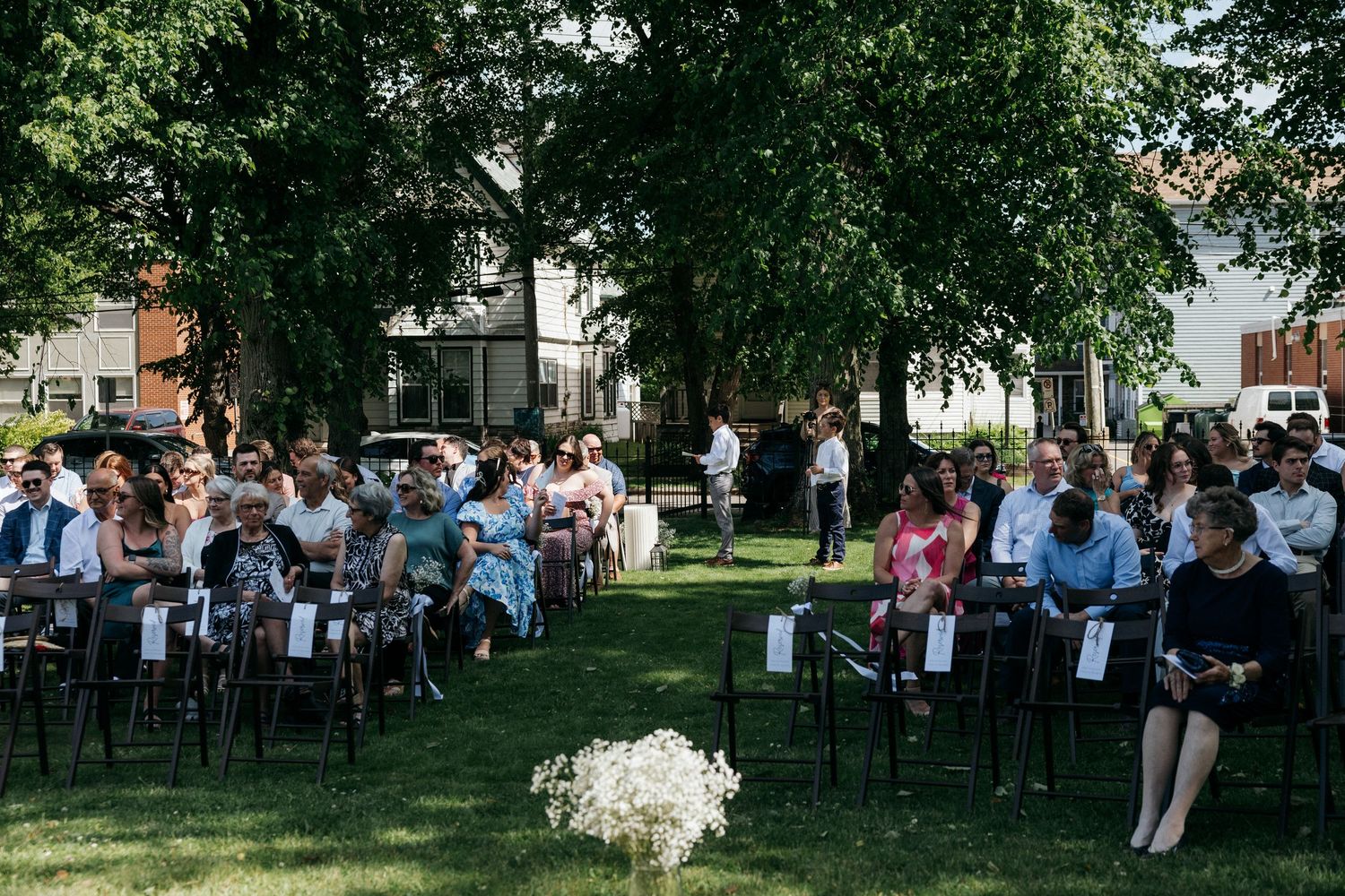Wedding guests seated on chairs arranged on a shaded lawn for an outdoor ceremony.