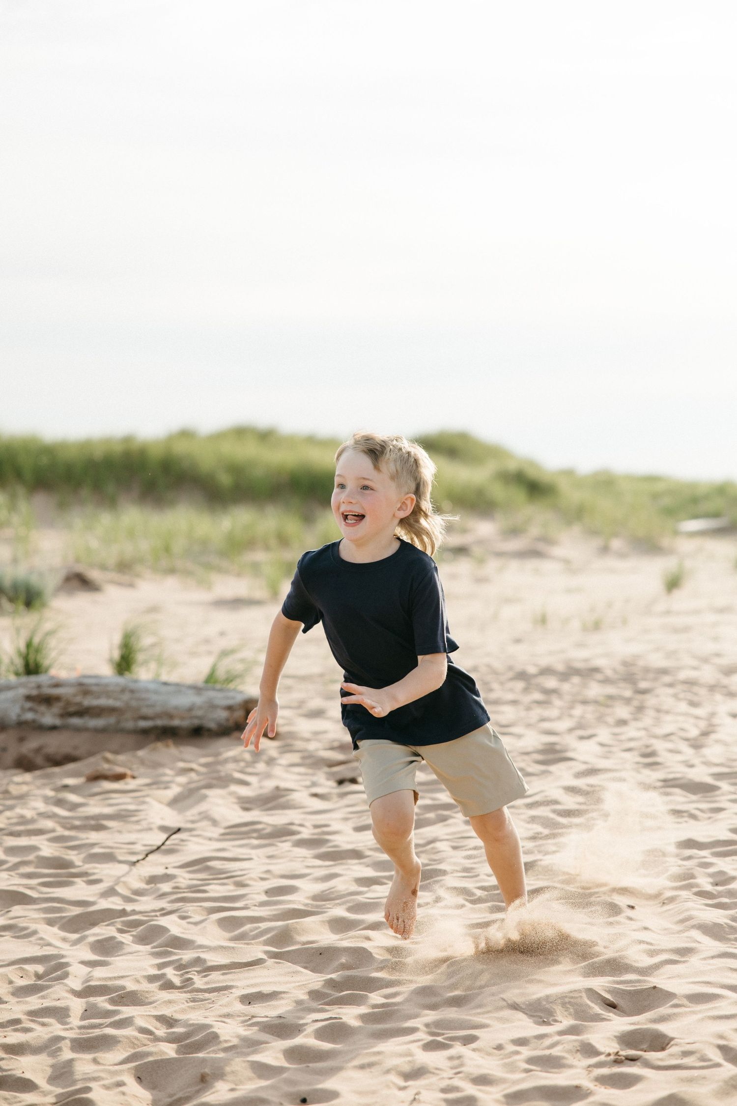 A happy toddler runs barefoot on sandy beach dunes wearing a black shirt and khaki shorts on a sunny day.