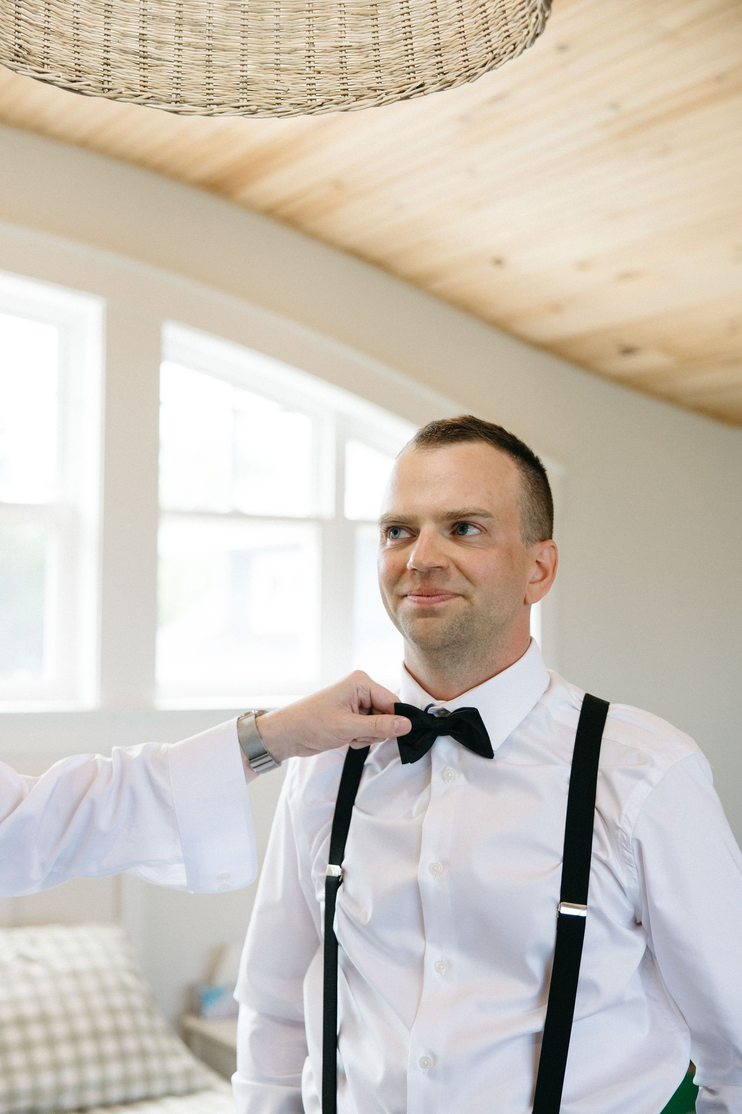 Groom in white shirt with black bow tie and suspenders being helped to dress before wedding ceremony.