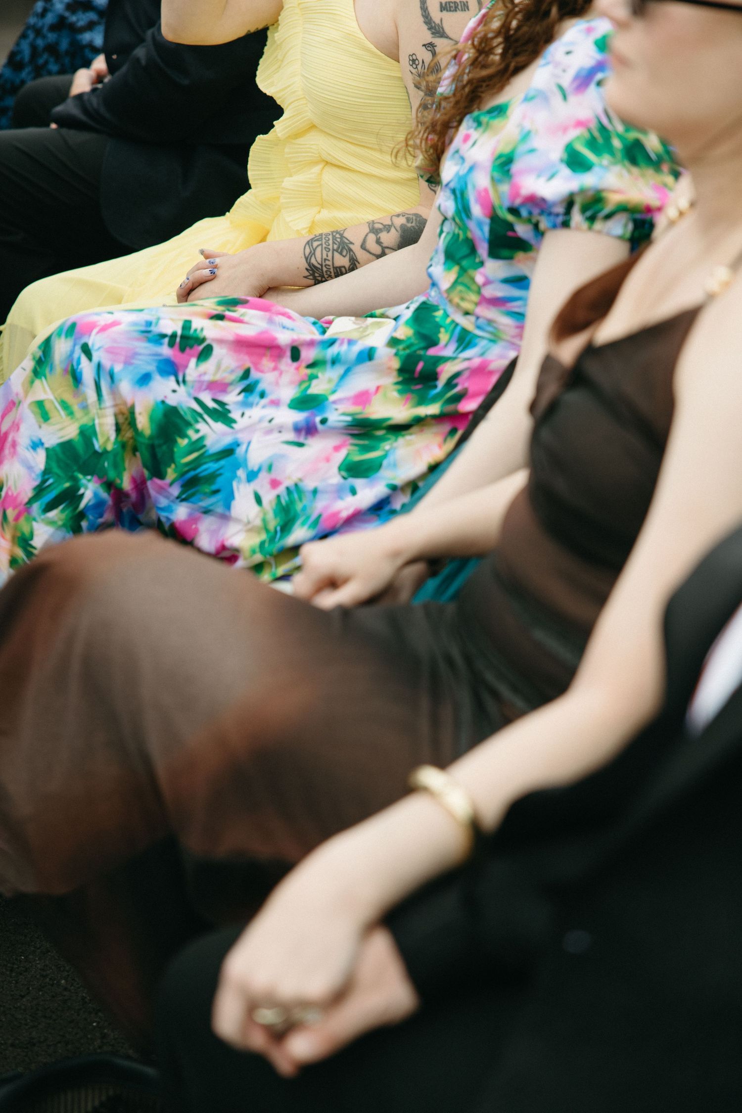 Wedding guests seated at ceremony wearing colorful floral and yellow dresses, with visible tattoos on one guest.