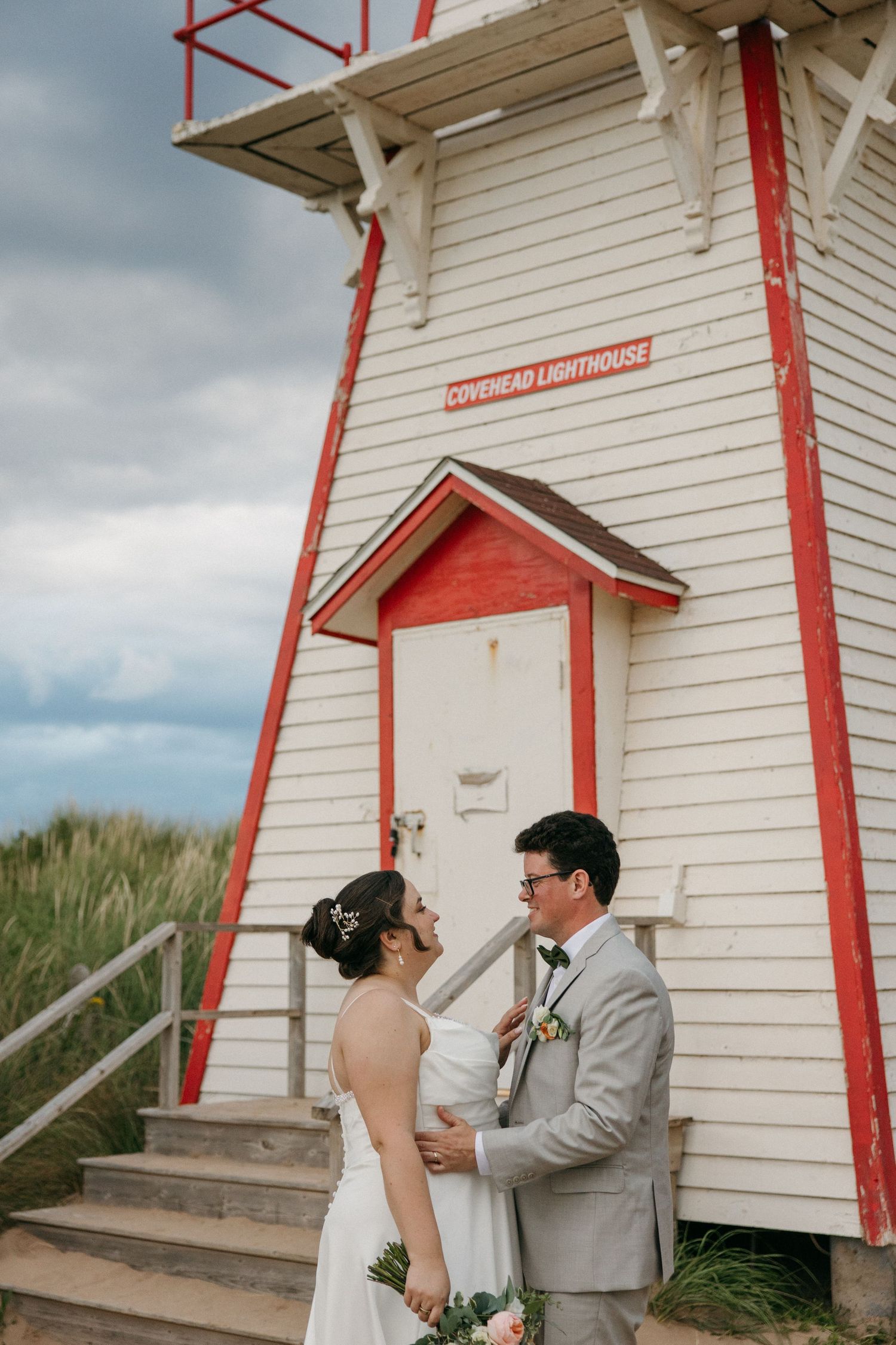 Bride and groom share tender moment in front of red and white Covehead Lighthouse under cloudy sky.