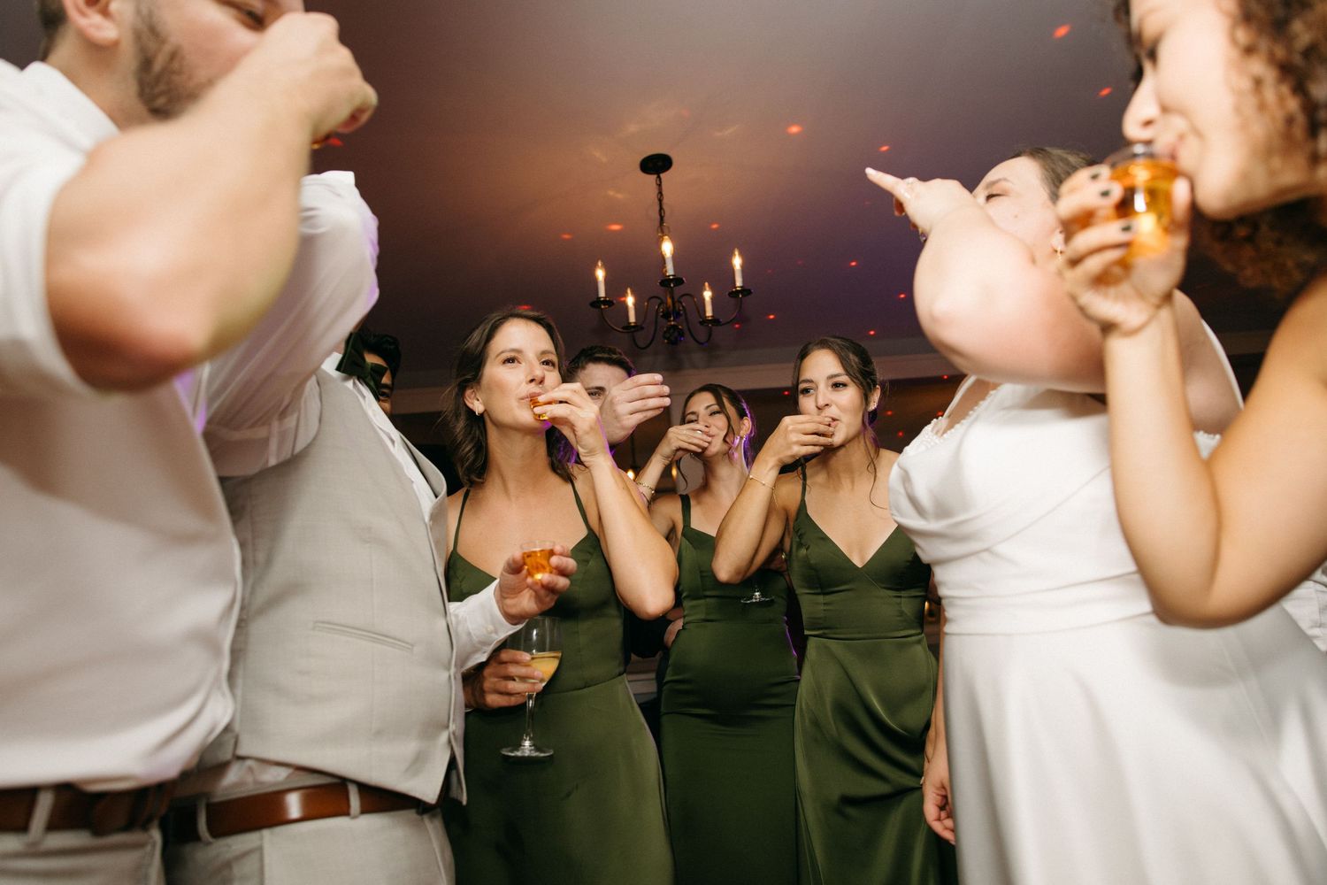 Bridesmaids in olive green dresses drinking shots with the bride on the wedding dance floor.