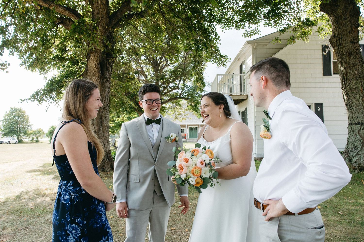 Bride with bouquet and groom in gray suit chat with guests under trees at outdoor wedding.