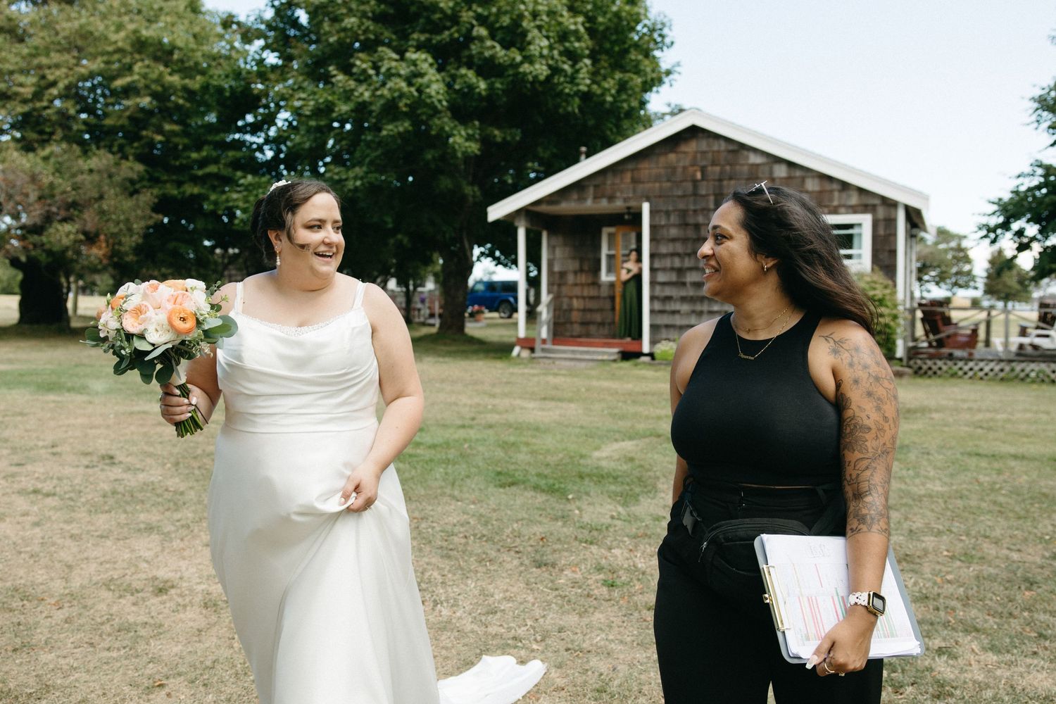 Bride in white gown holding peach bouquet shares a joyful moment with wedding coordinator outdoors.
