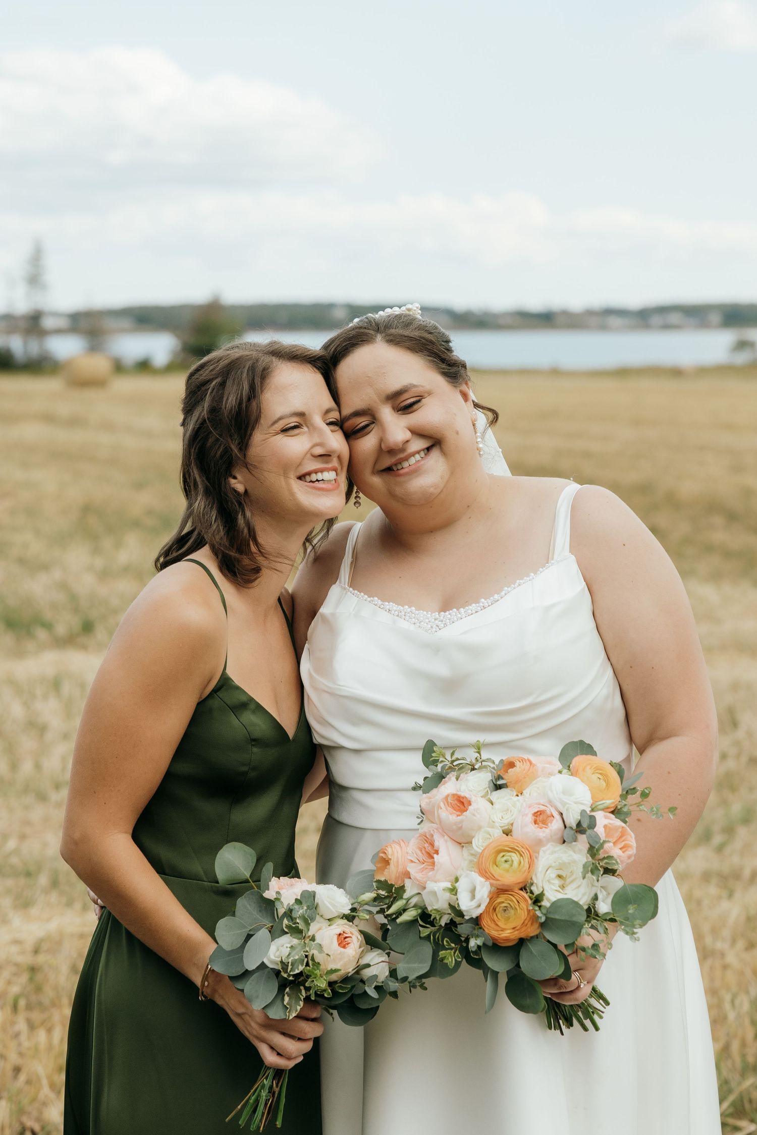 Bride in white gown and bridesmaid in green dress share a joyful embrace in a golden field.