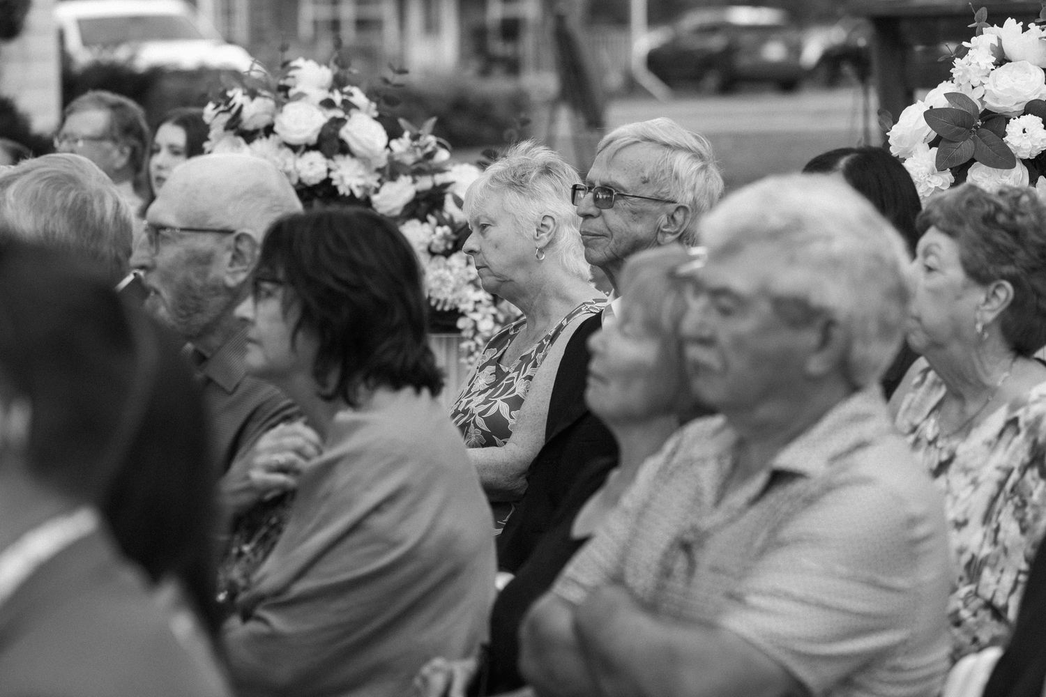 Black and white photo of wedding guests seated at an outdoor ceremony with floral hair accessories.
