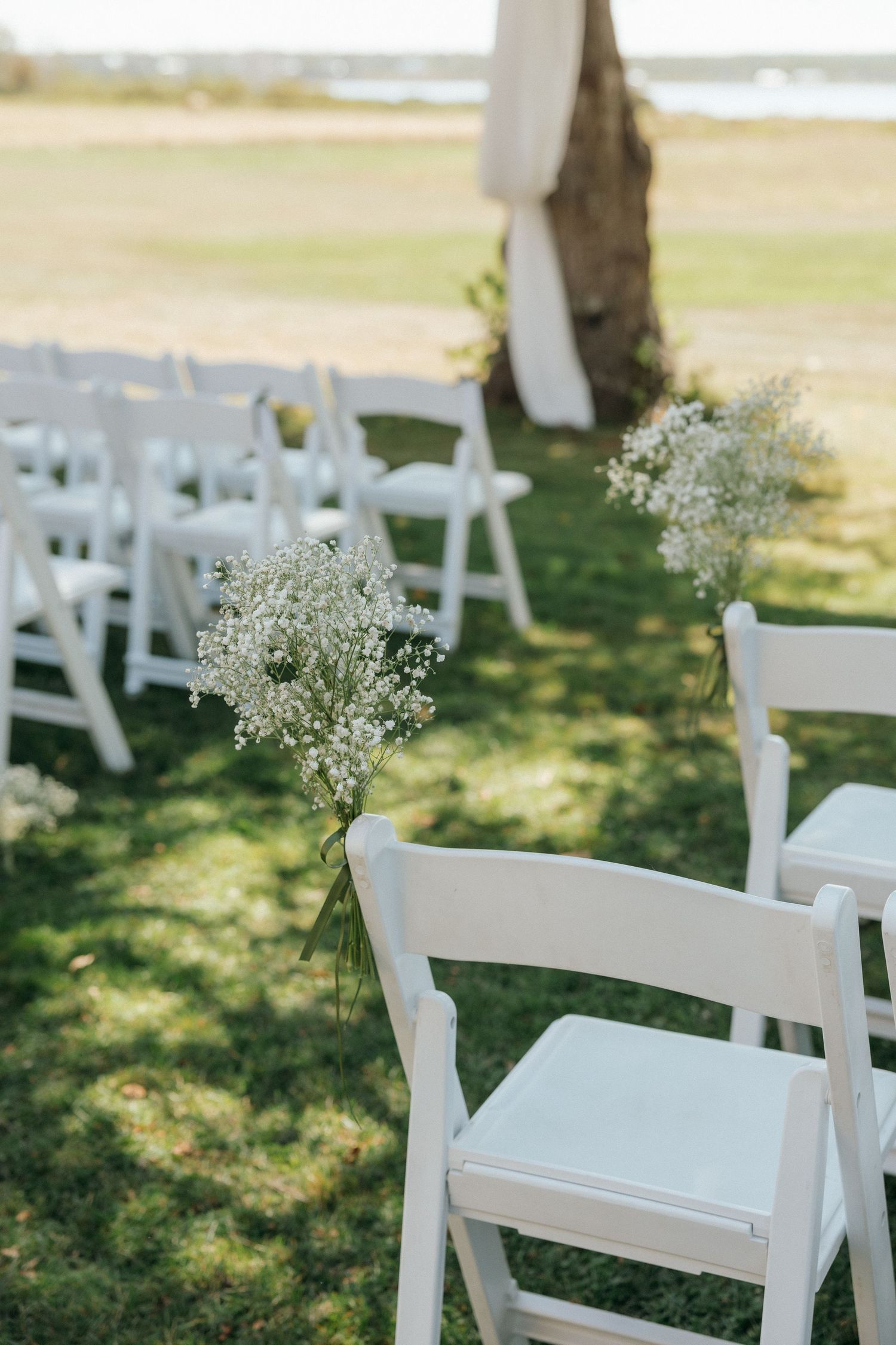 White folding chairs decorated with baby's breath line a sunny outdoor wedding ceremony aisle.