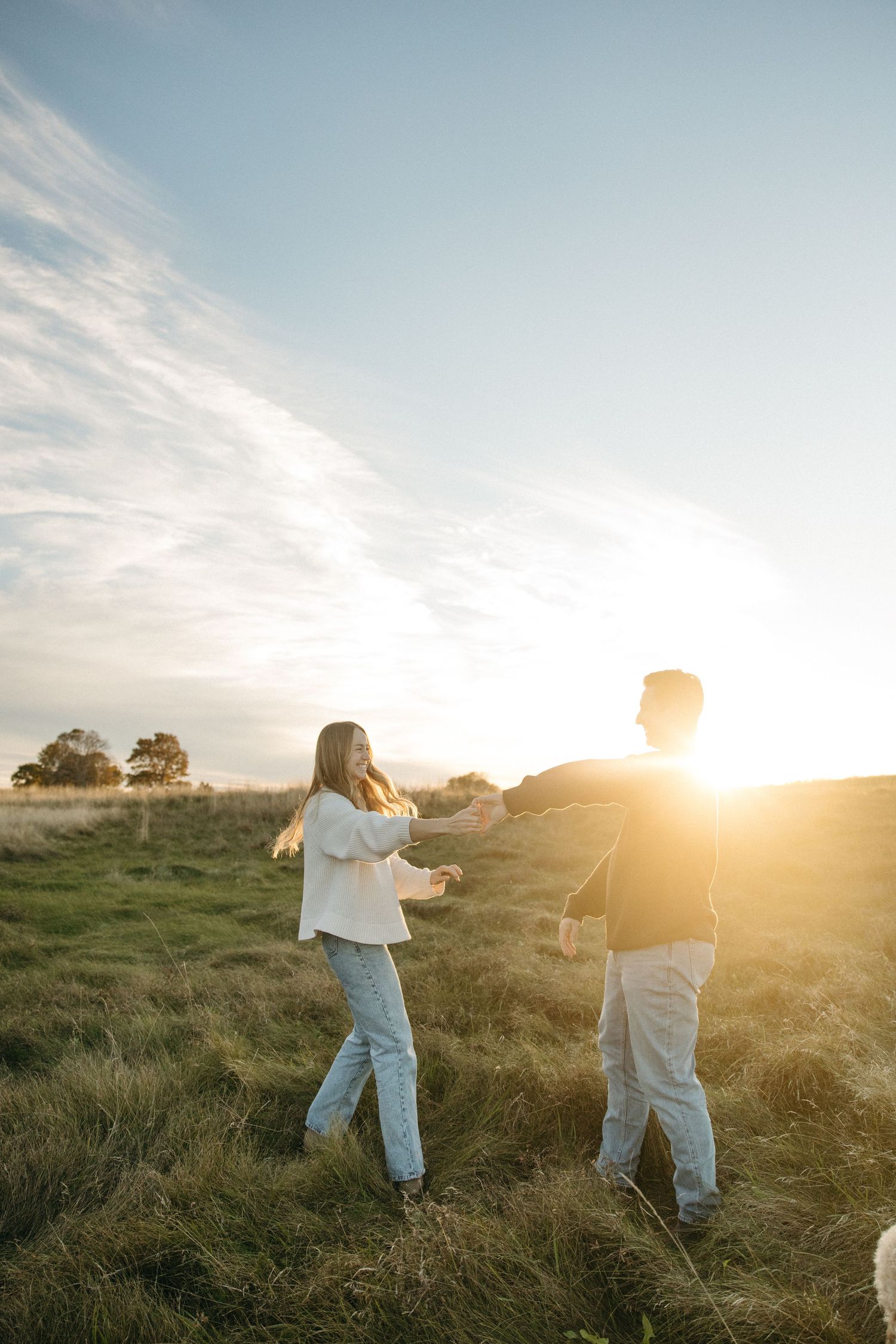 Couple holding hands and dancing in a sunlit grassy meadow as the golden sunset glows behind them.