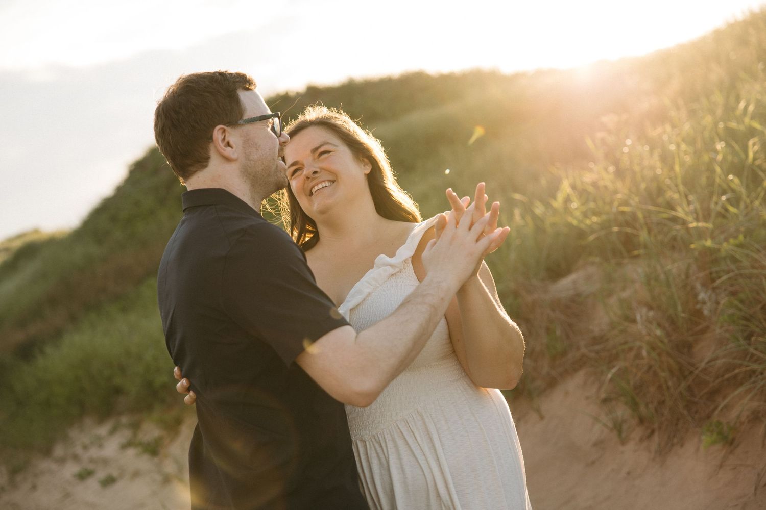 Romantic couple embracing in warm golden sunset light among coastal grass dunes.