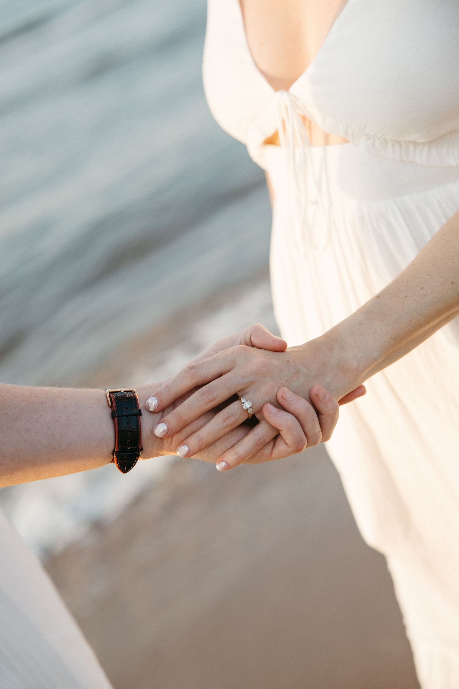 Close-up of couple holding hands near ocean shore, woman wearing diamond engagement ring.