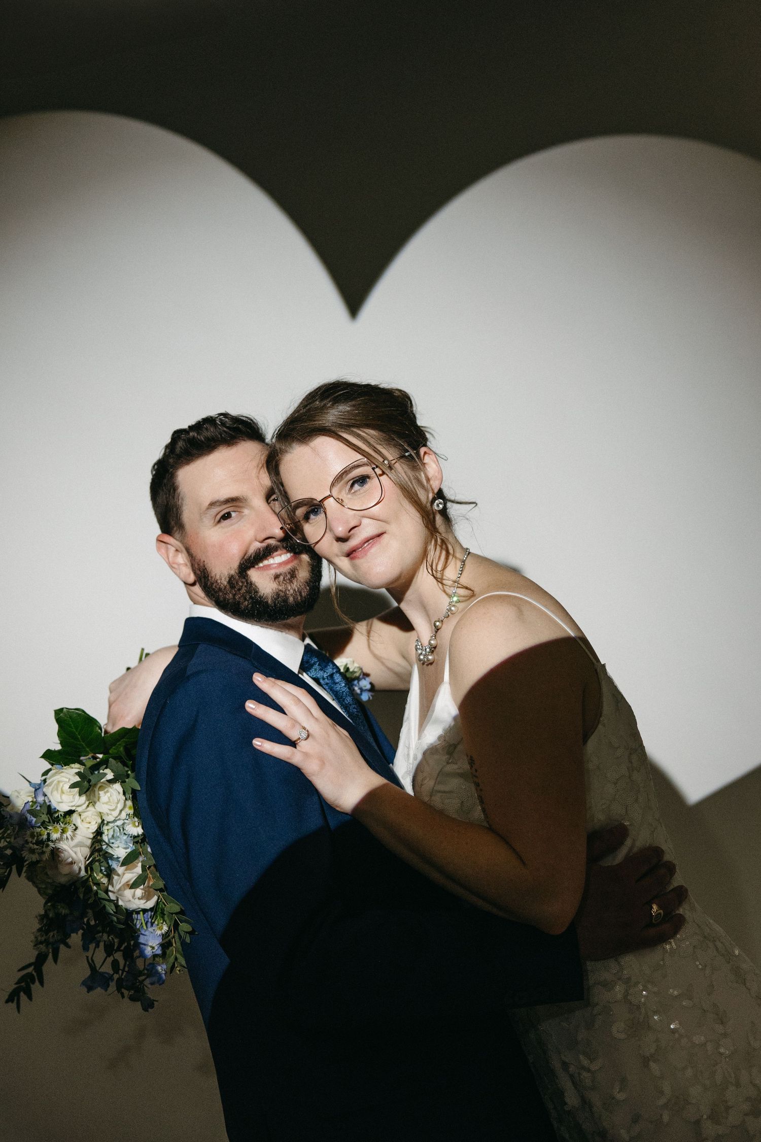 A smiling bride and groom embrace in front of a heart-shaped light projection on their wedding day.