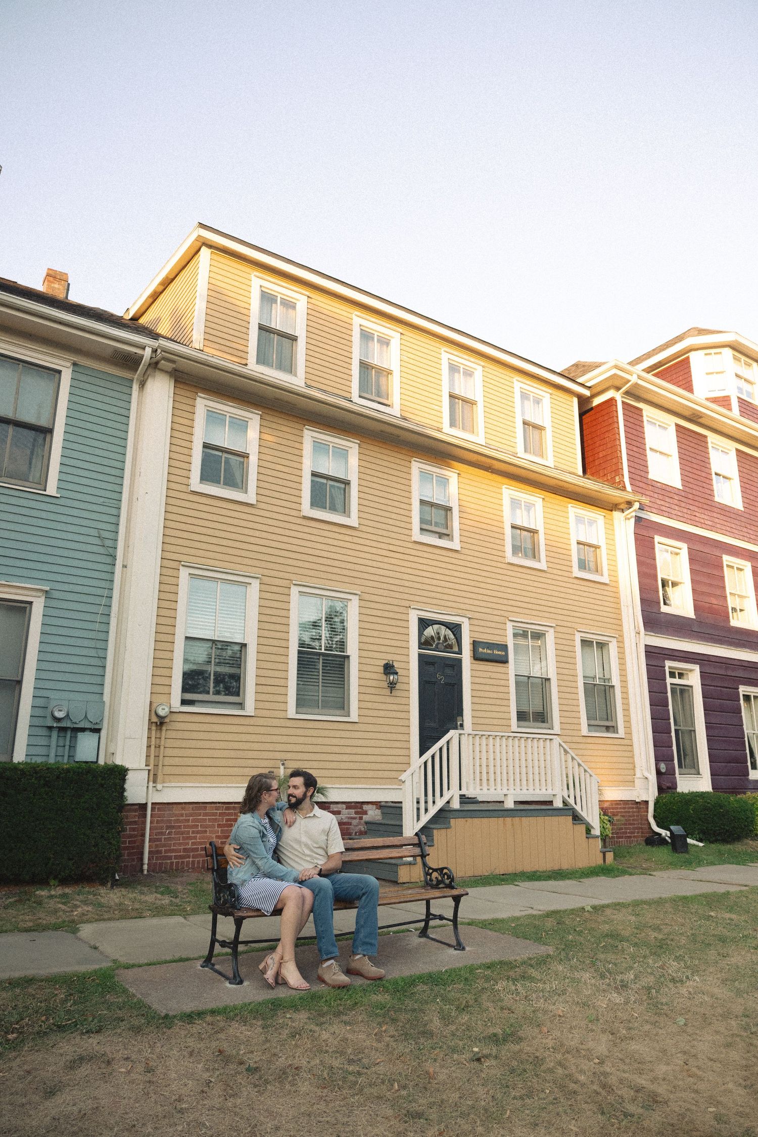 A couple relaxes on a bench in front of colorful row houses on a residential street.